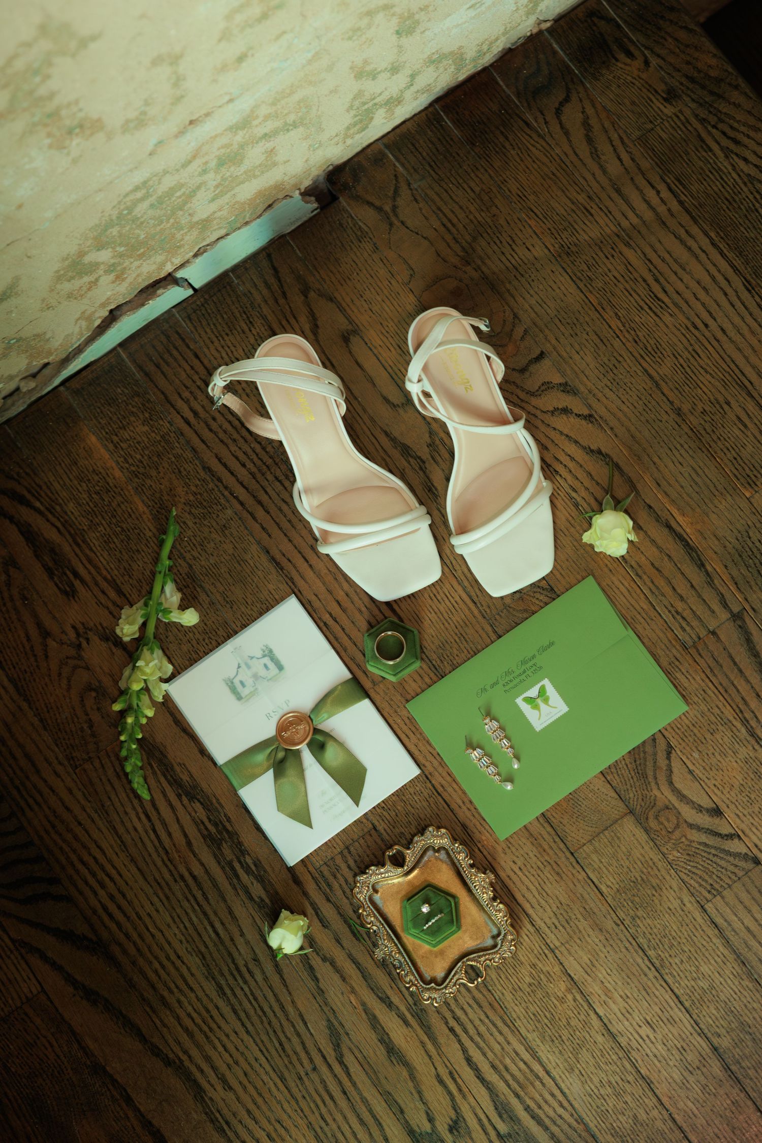 White block-heel wedding sandals displayed on wood floor with green stationery, wax seal, ring dish, pearls, and florals.