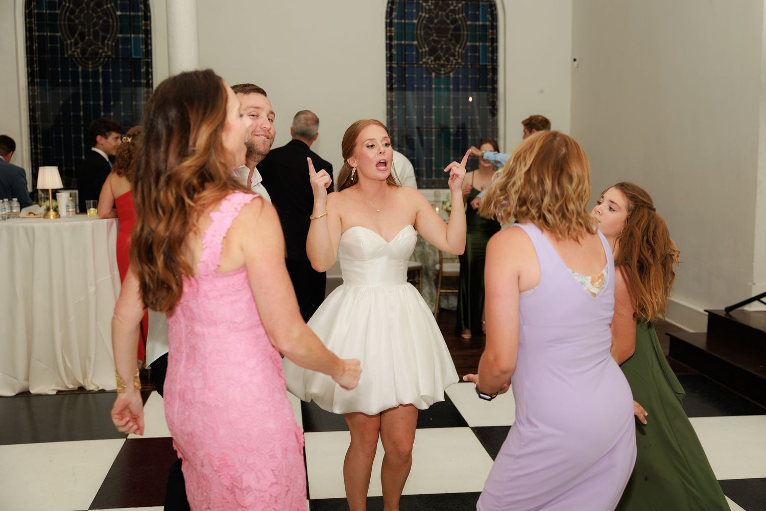 Bride in white dress dancing joyfully with bridesmaids in pink and lavender gowns on checkered dance floor at wedding reception.