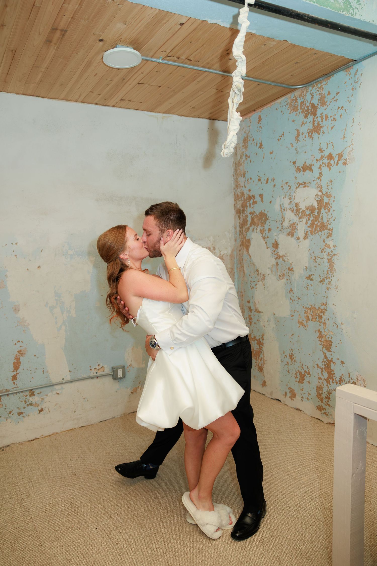 Couple sharing a romantic kiss in wedding attire, bride in white mini dress, groom in white shirt, against rustic peeling blue walls.