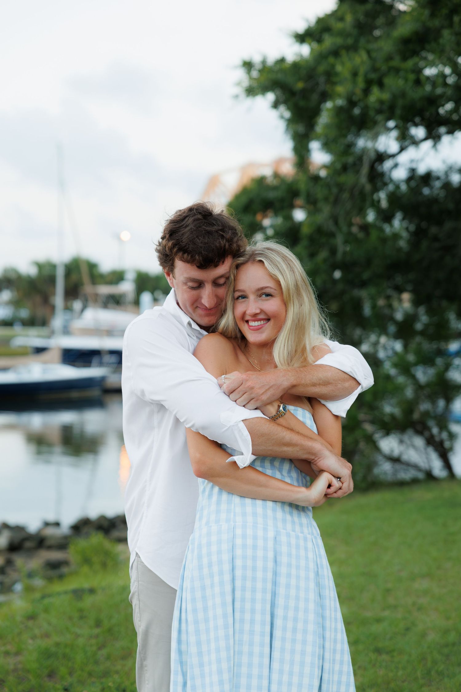 Couple embracing near a marina, woman in blue gingham dress smiling, man in white shirt hugging her from behind.