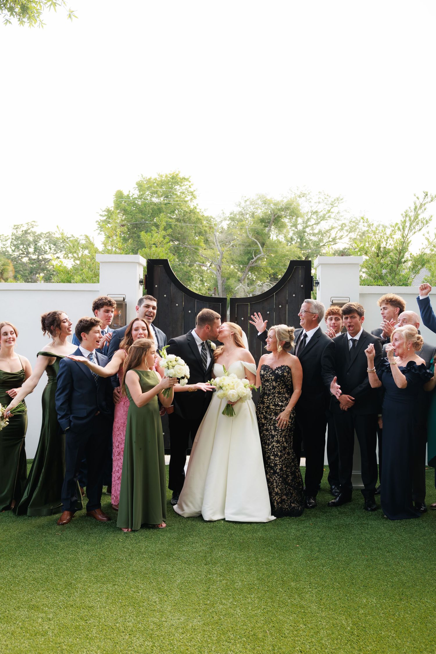 Wedding party celebrating with bride and groom kissing, bridesmaids in olive green gowns, black iron gate backdrop.