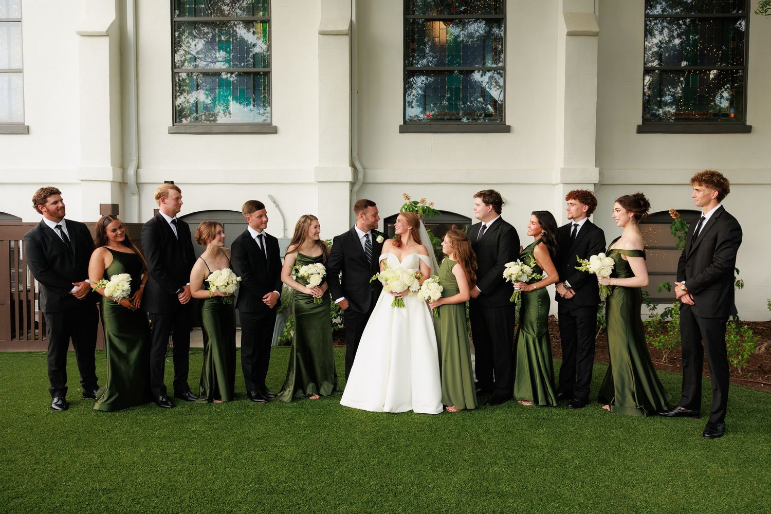 Wedding party in green bridesmaid dresses and black suits posing on lawn before white church.