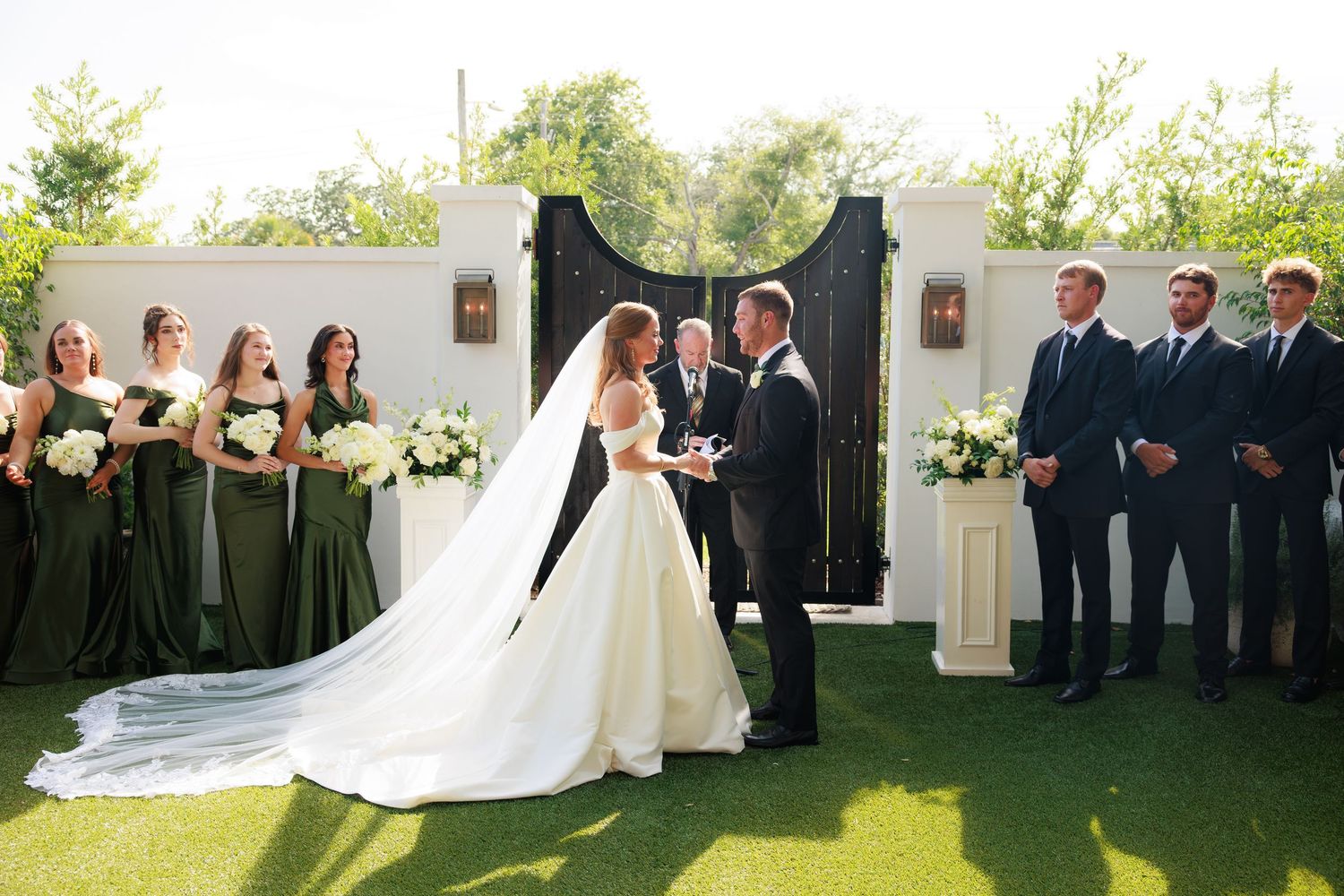 Outdoor wedding ceremony with bride in white gown, groom, officiant, bridesmaids in green dresses, and groomsmen in dark suits.