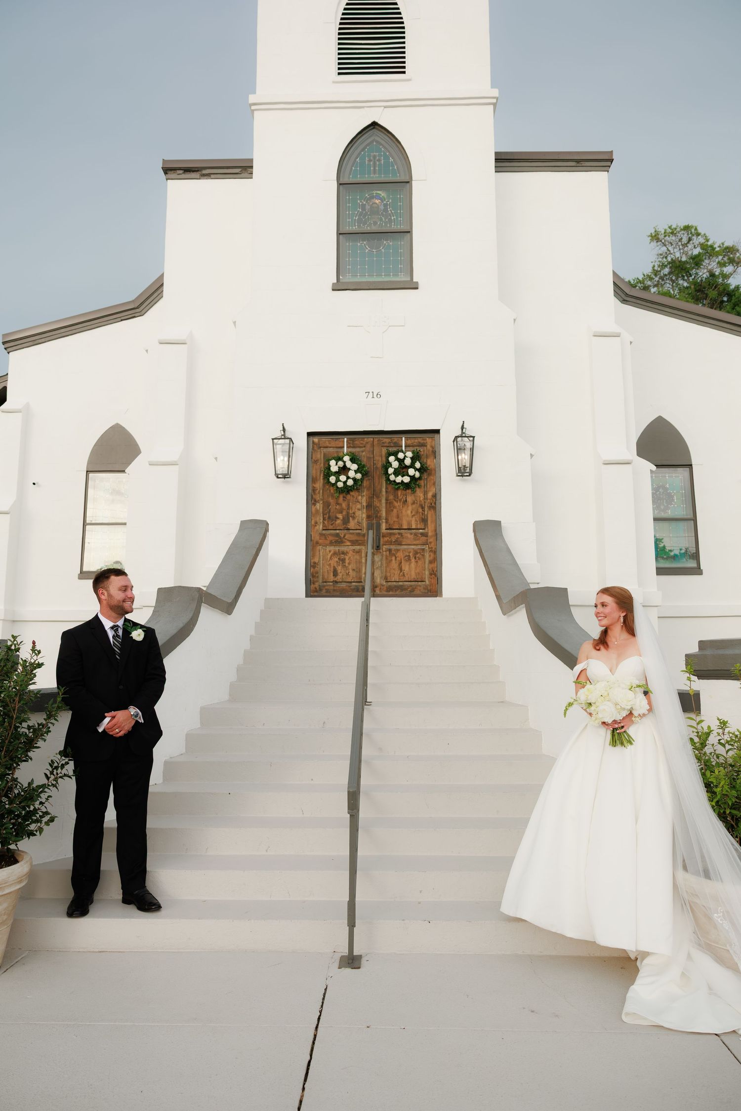 Bride and groom share a first look moment on steps of a white church with wooden doors and wreaths.