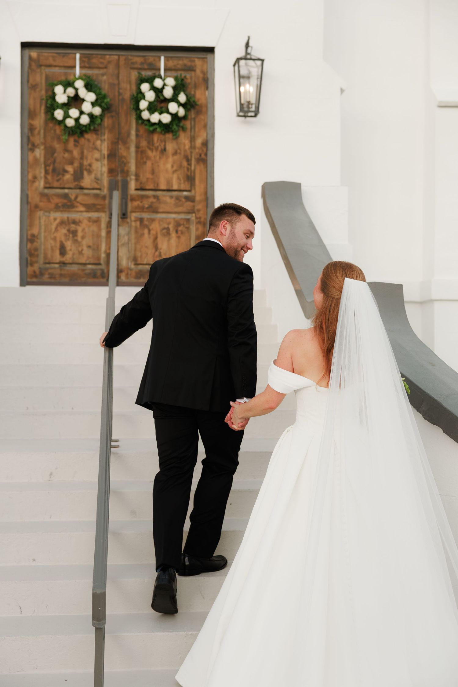 Bride and groom walking hand-in-hand up white stairs toward wooden doors with floral wreaths.