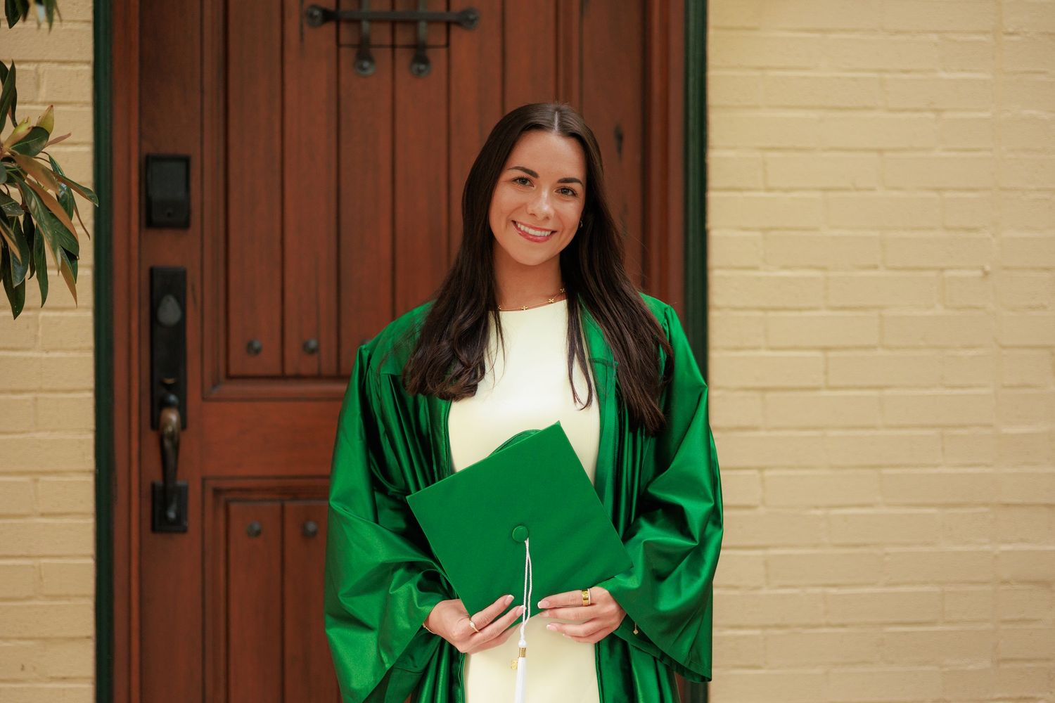 Smiling woman in green graduation gown and cap standing in front of a wooden door.