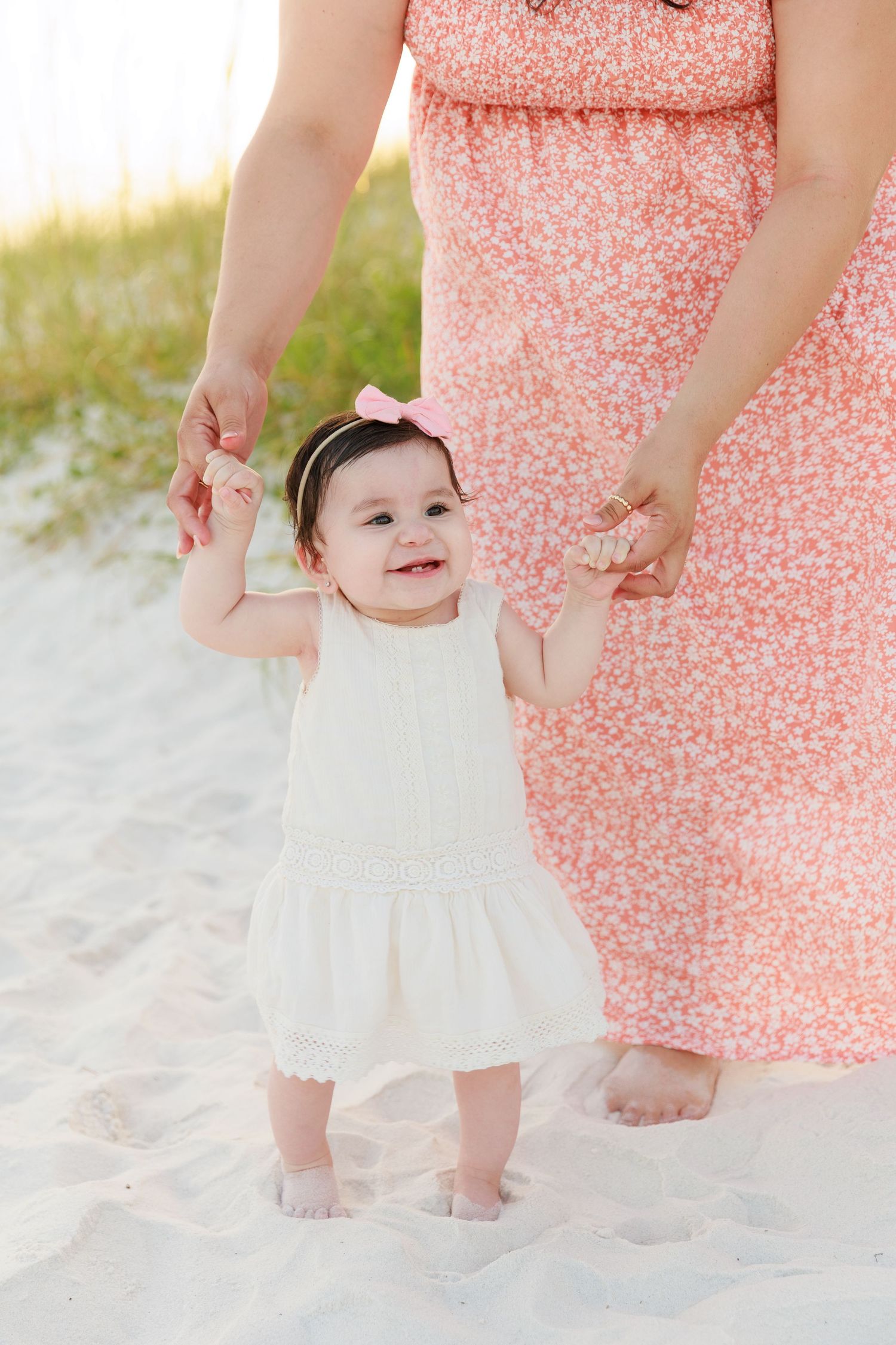 Smiling baby girl in white dress holds mother's hands on sandy beach, wearing pink bow headband.