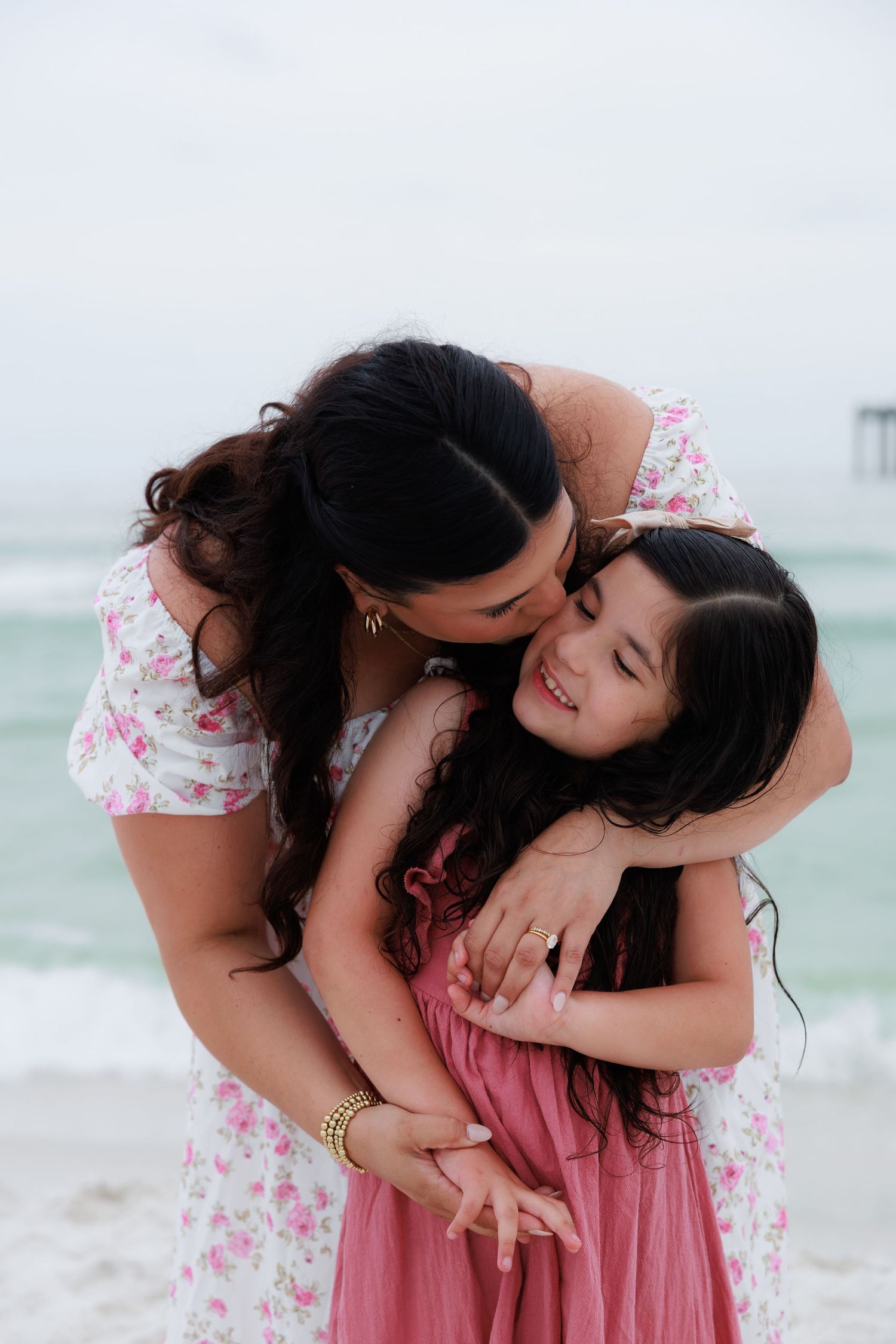 Mother embracing and kissing smiling daughter with long dark hair on beach, both wearing pink and white dresses.