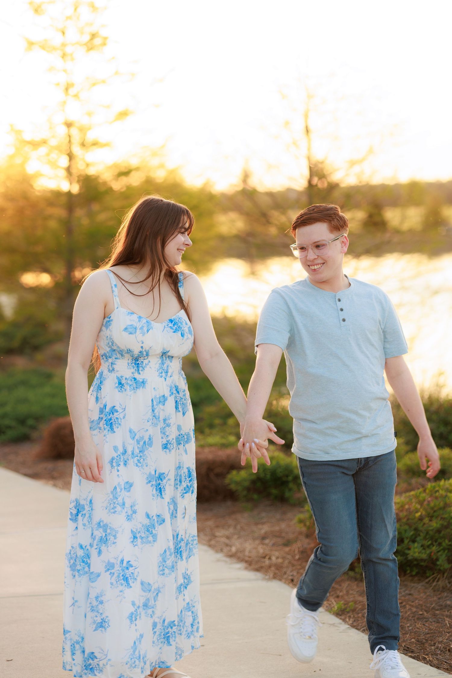 A young couple holds hands and smiles while walking along a path near a lake at golden hour sunset.