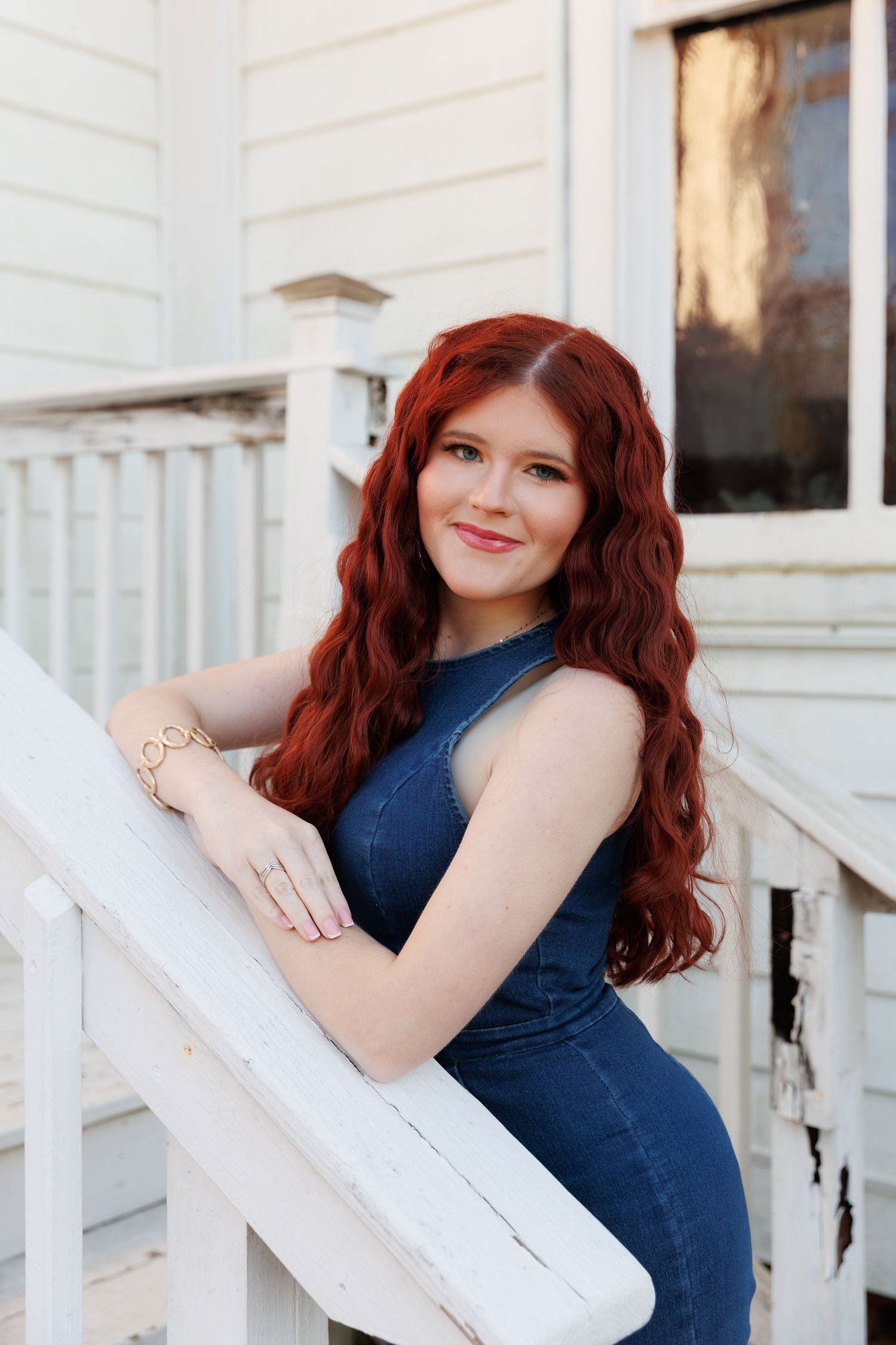 Woman with vibrant red curly hair poses against white porch railing in navy blue dress.
