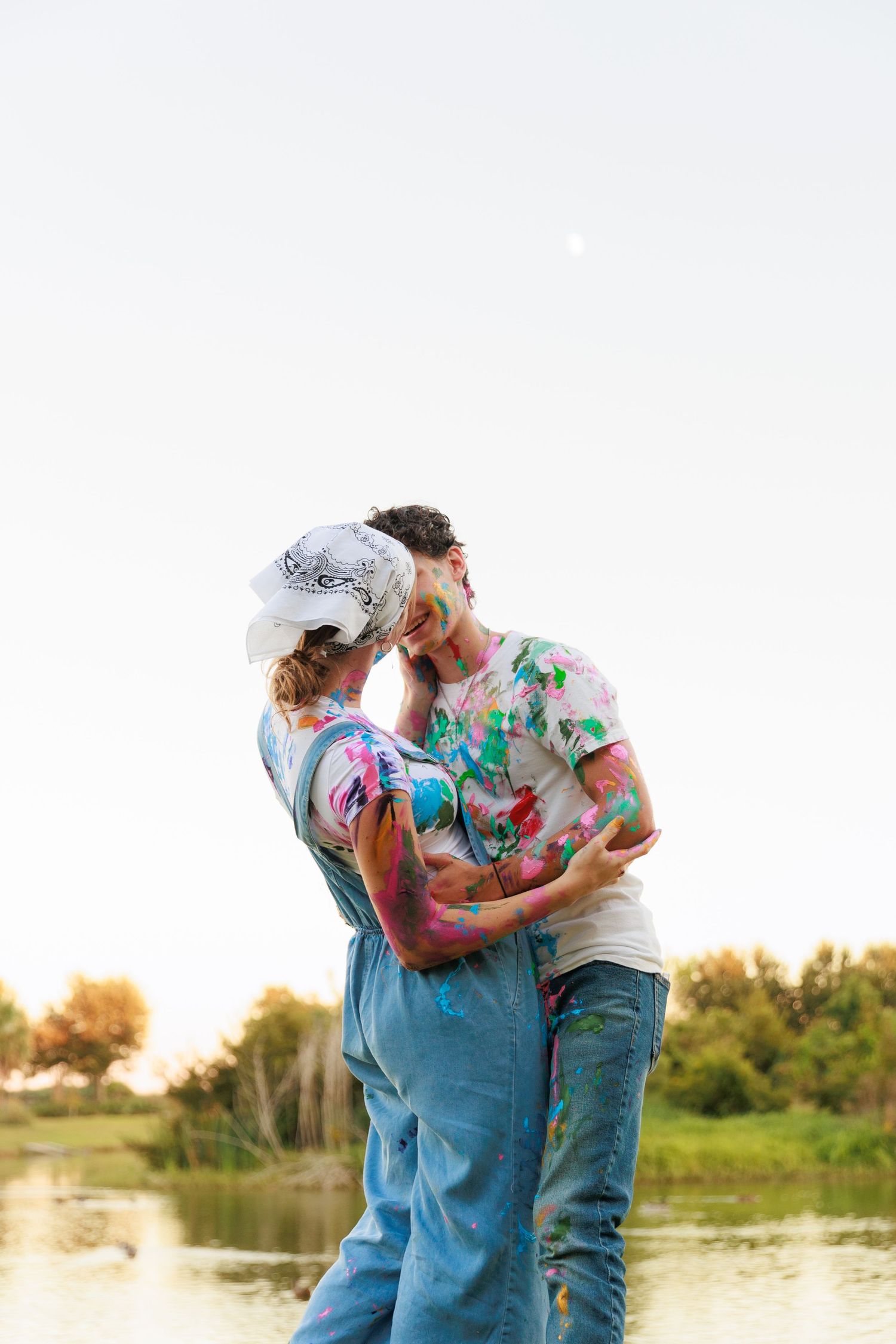 A couple covered in colorful paint powder embraces by a lake at sunset during a Holi celebration.