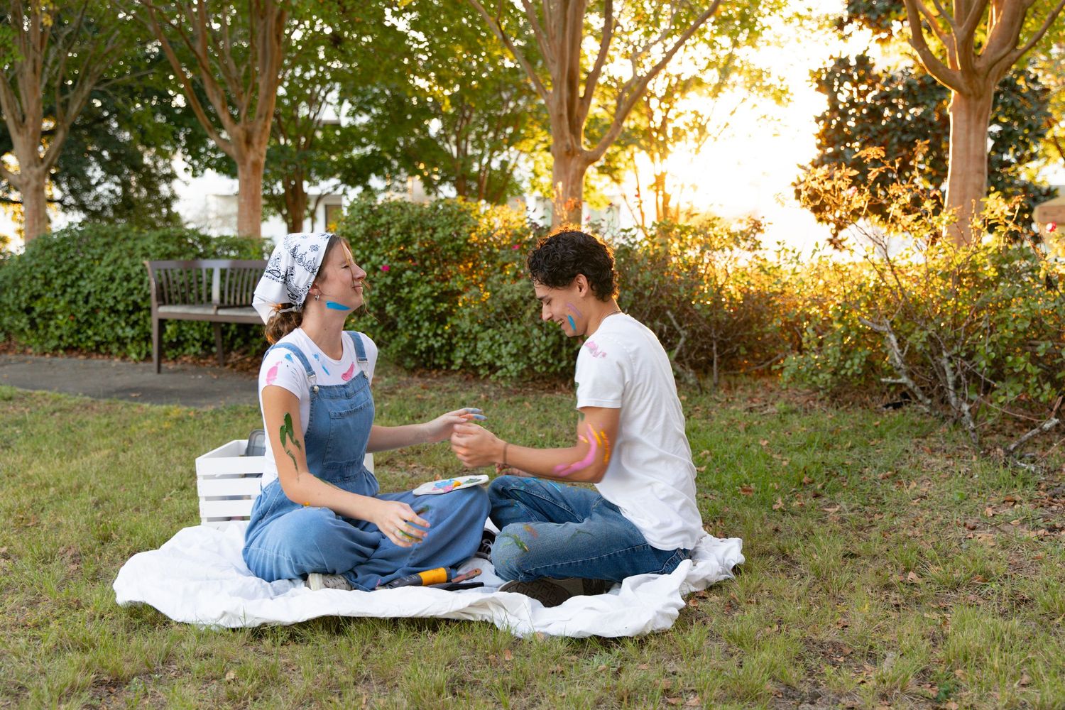 Two people sit on a white blanket in a grassy outdoor setting during golden hour.