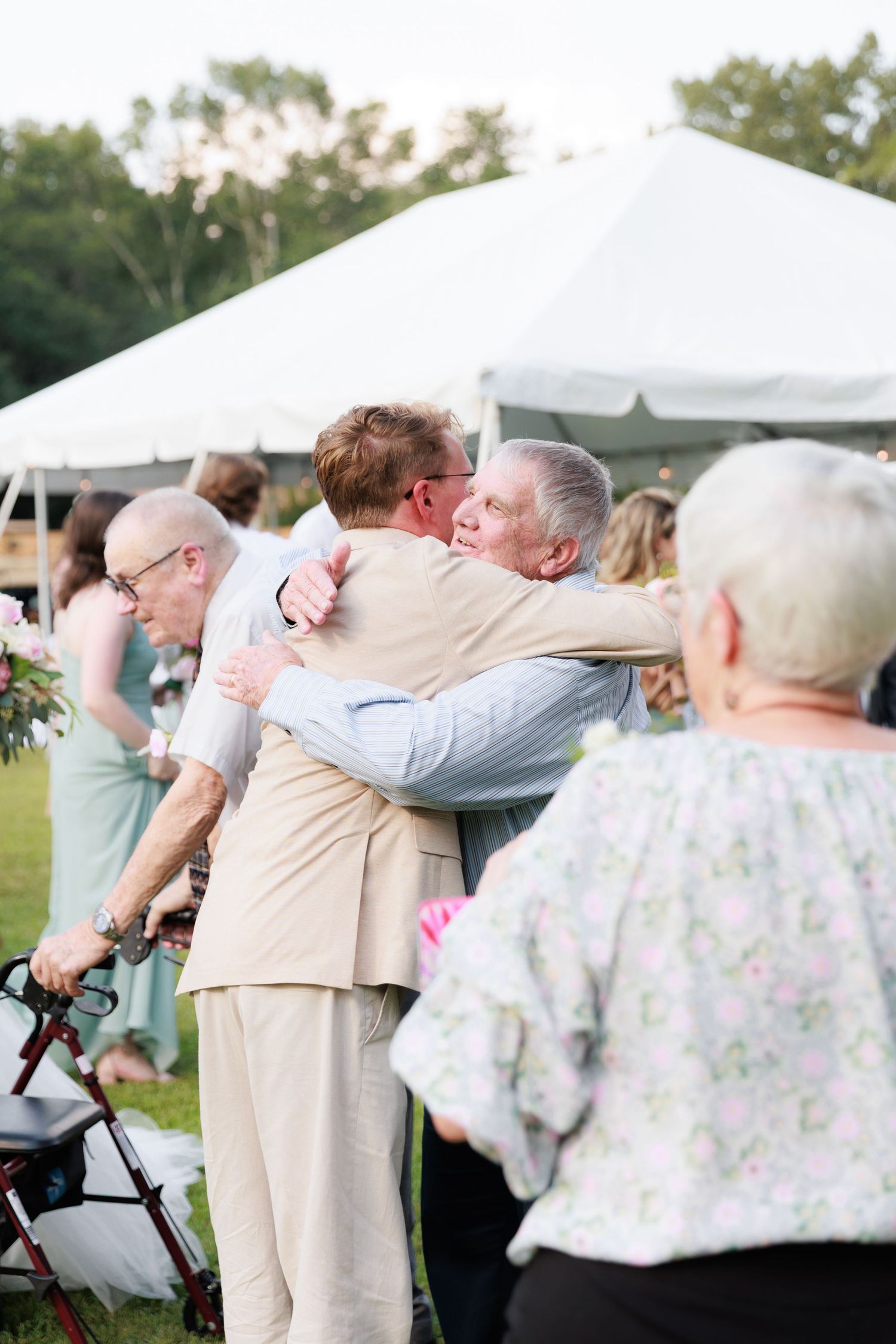 Wedding guests shield their eyes from the sun while watching an outdoor ceremony at a tented venue.