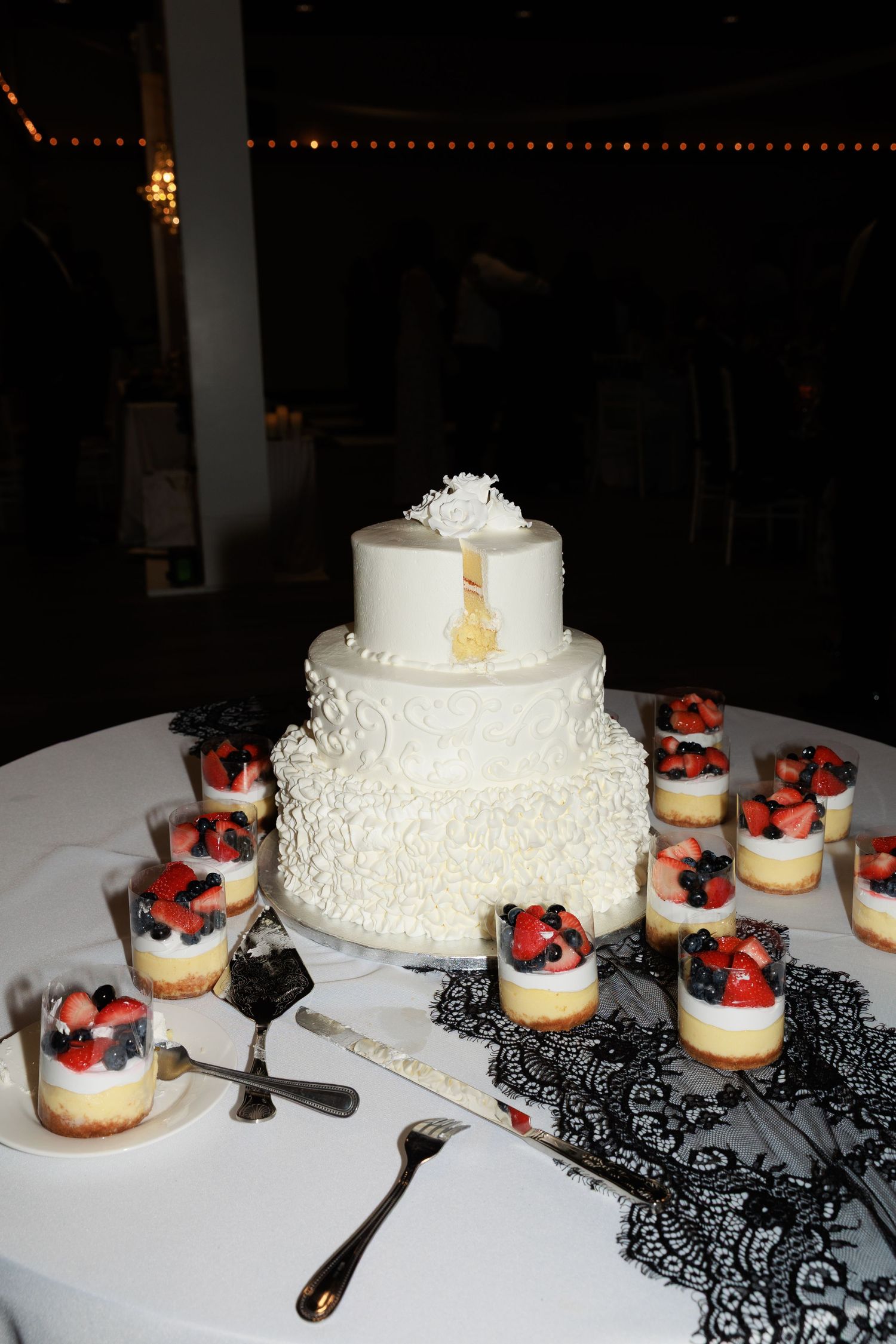 White wedding cake with lace details displayed alongside mini desserts decorated with red and black accents on a table.