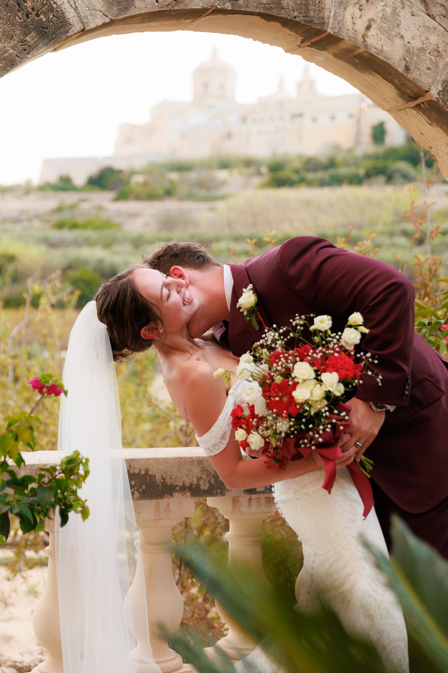 A romantic wedding couple shares an intimate moment against a stone archway with red and white floral accents.