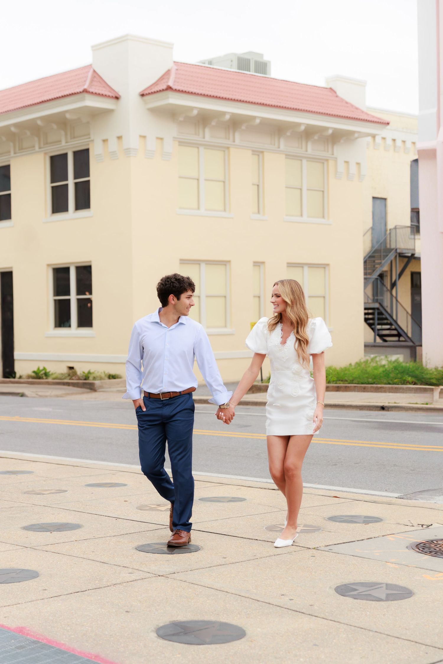 A couple walks hand in hand on a sidewalk in front of a cream-colored building with a pink roof.