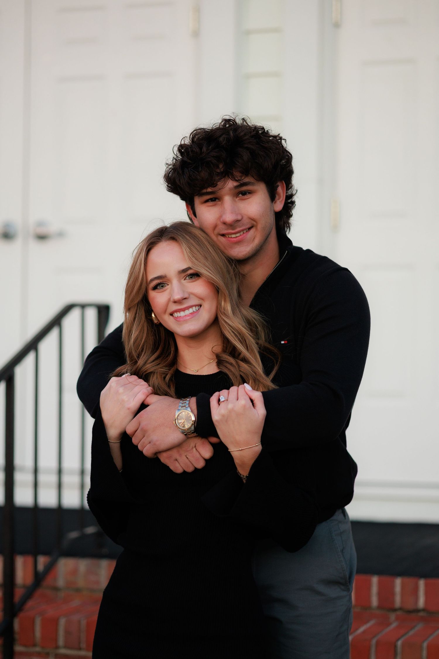 A couple in black outfits embraces while standing in front of a white building with stairs.