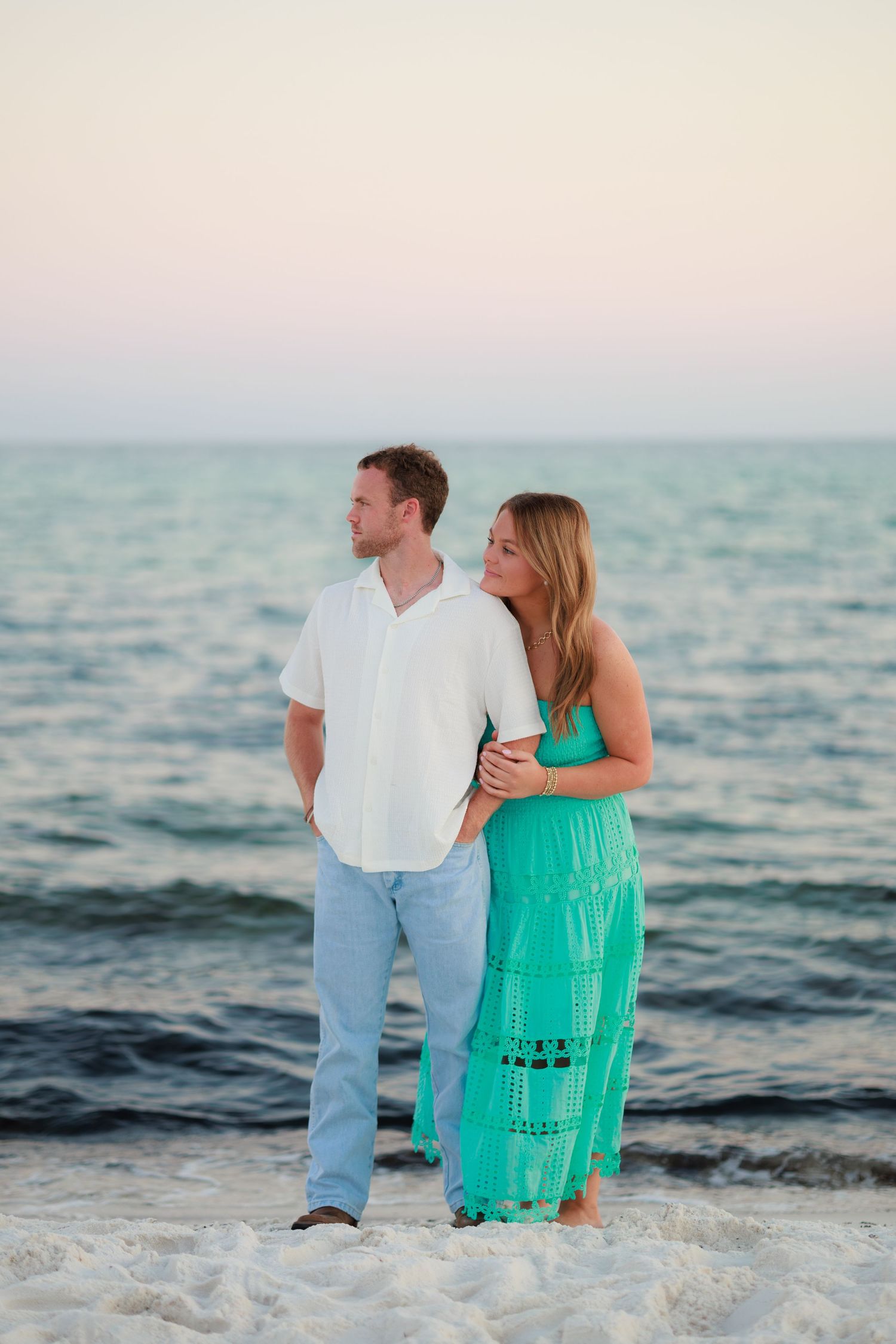 Couple in white shirt and turquoise dress stands together on beach at sunset with ocean waves behind them.