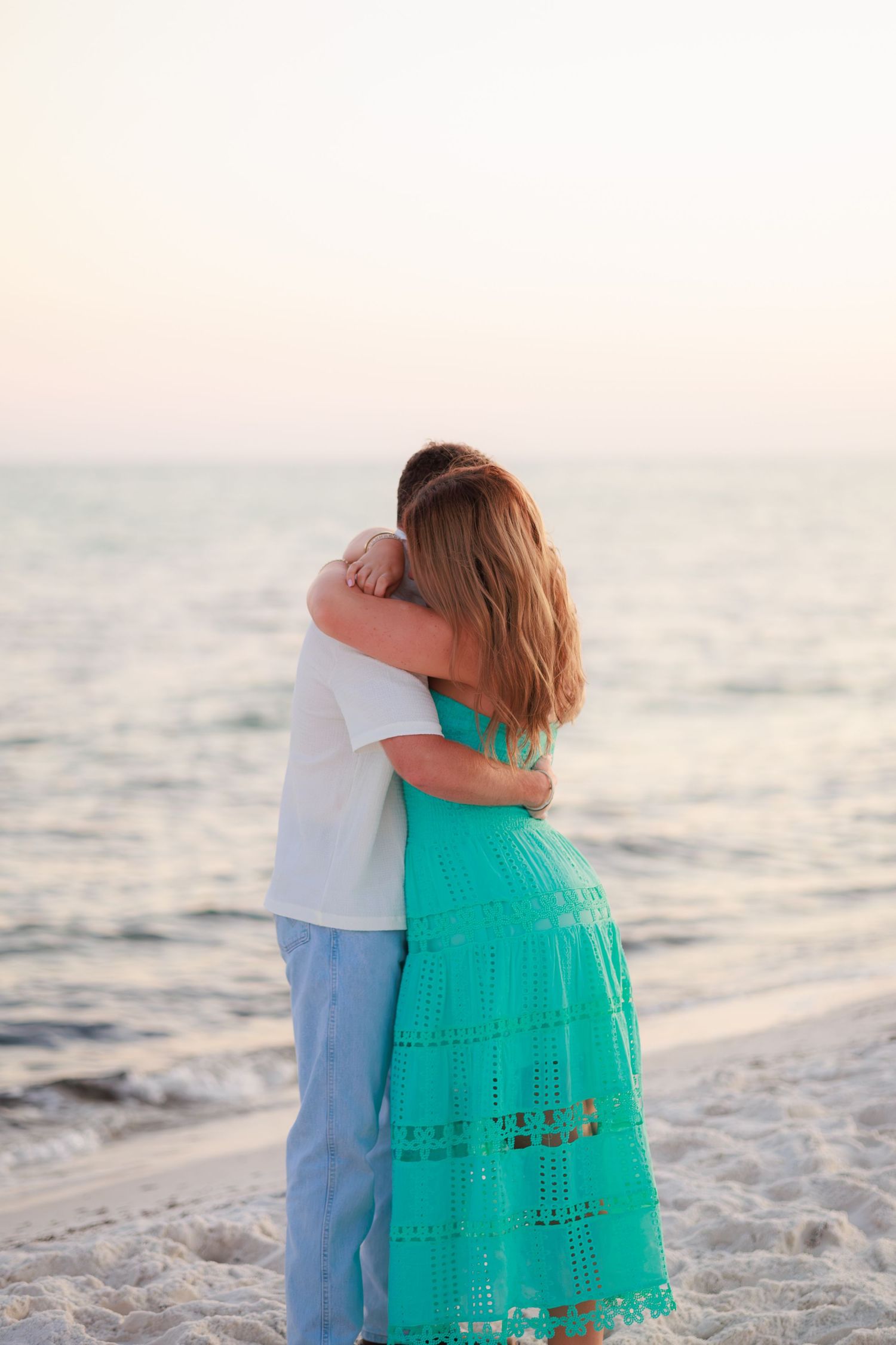A romantic couple embraces on a sandy beach at dusk with the ocean waves behind them.