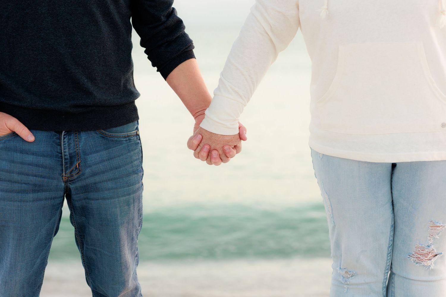 A couple wearing casual clothing holds hands while walking together on a beach.