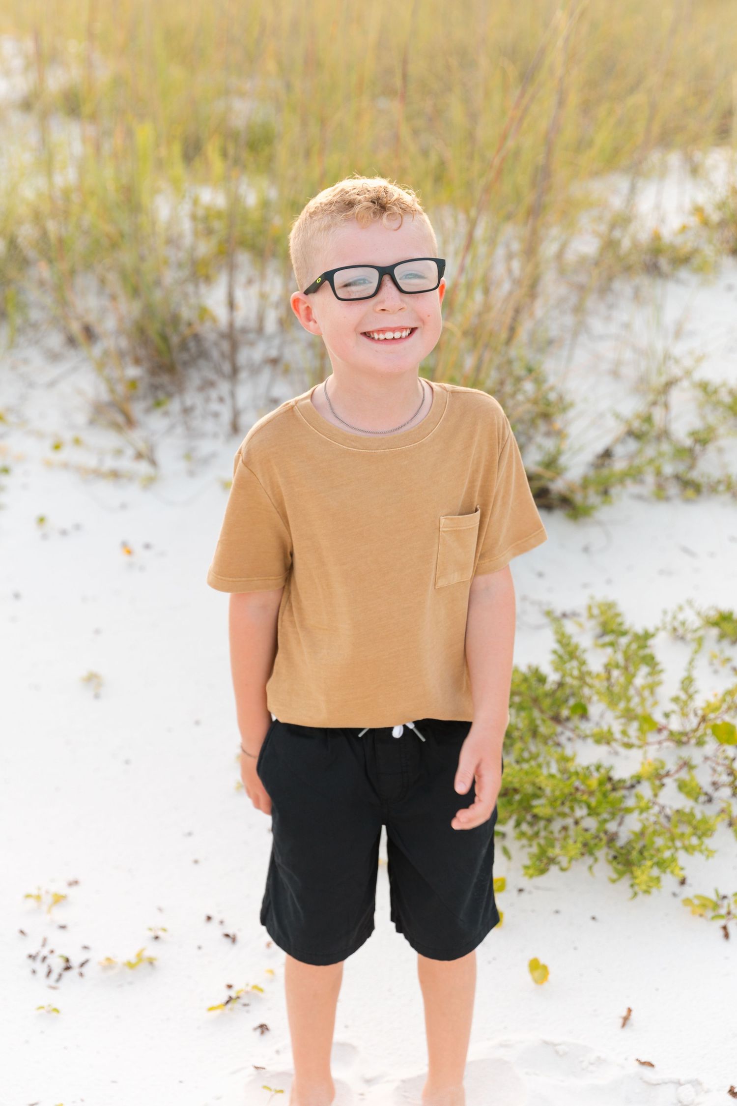 Child in casual attire stands smiling on white sand with coastal vegetation.