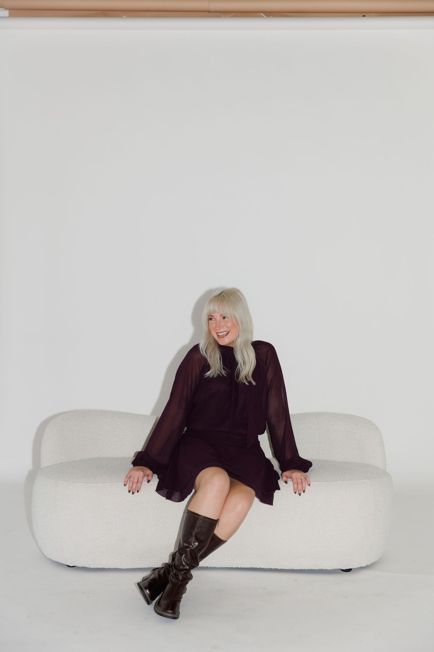 Stylish figure in dark clothing poses on curved white contemporary seating against stark white backdrop.