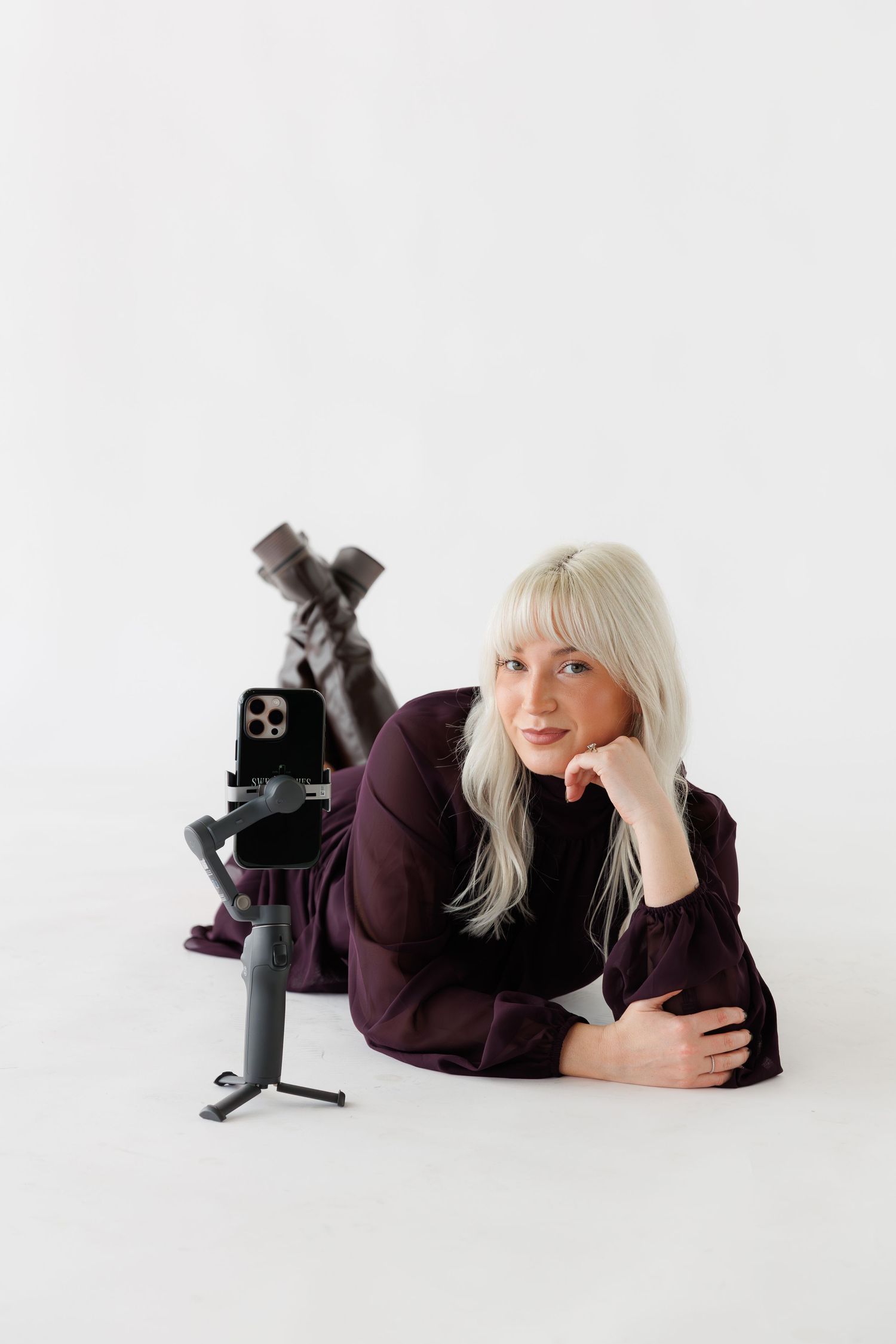 Person in burgundy sweater lies on white floor with camera equipment in minimalist studio.