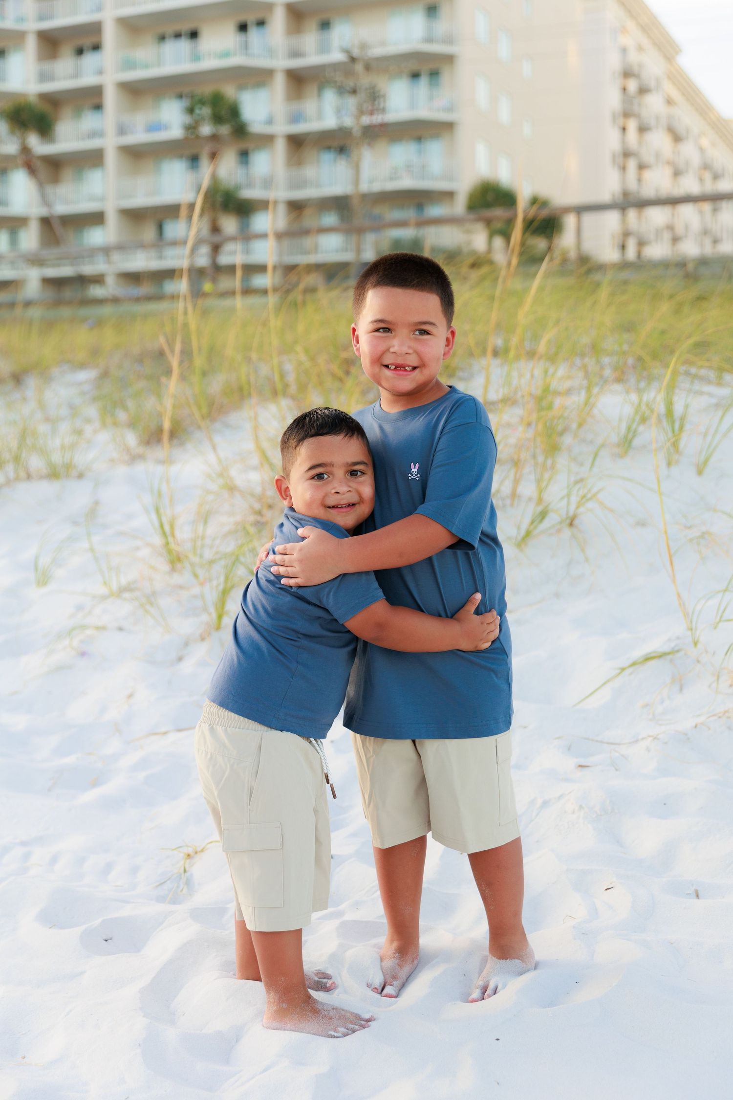 Two children in matching blue shirts and khaki shorts embrace while standing on white sand beach with condos in background.