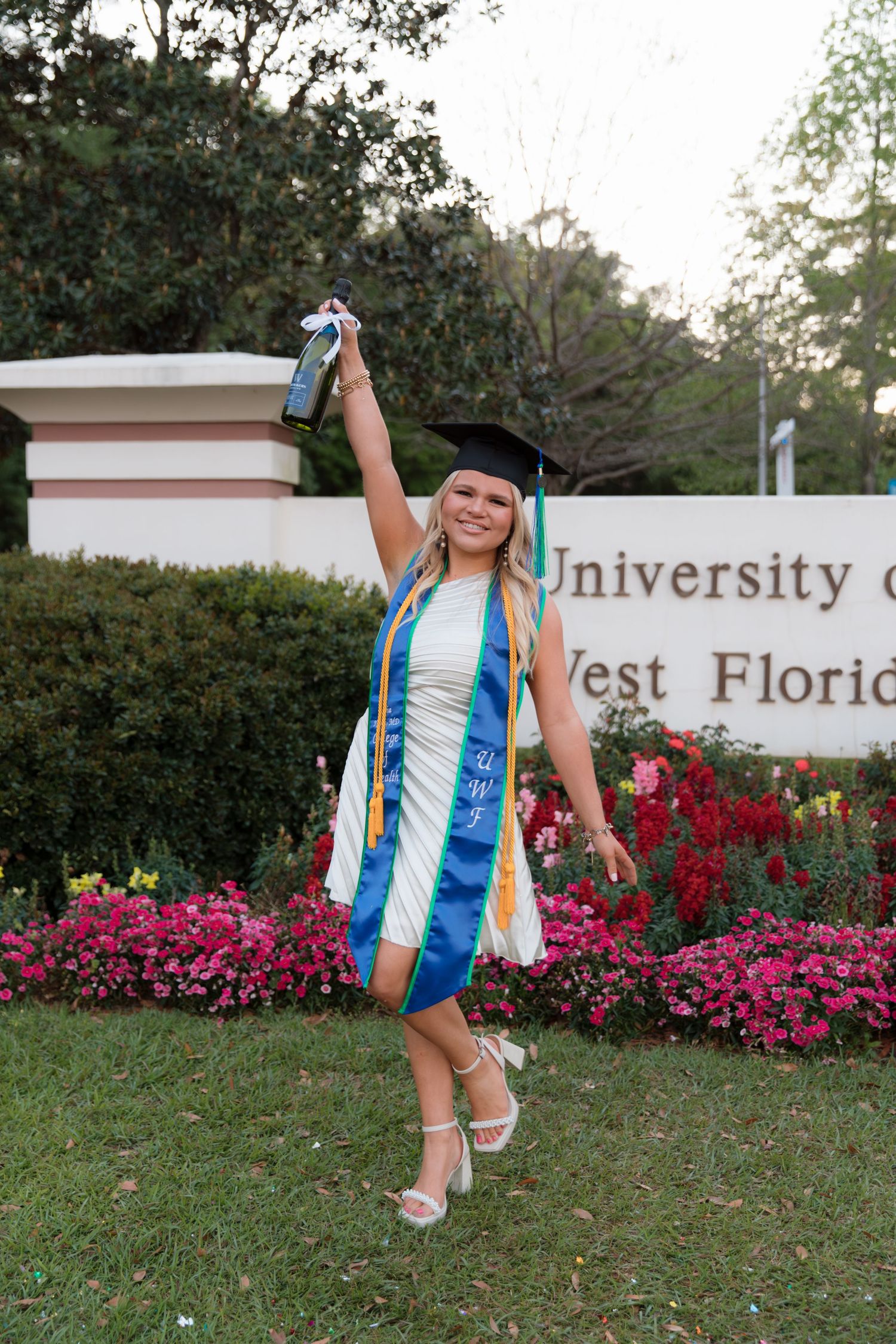 Graduate celebrates near university sign surrounded by vibrant pink flowers and greenery.
