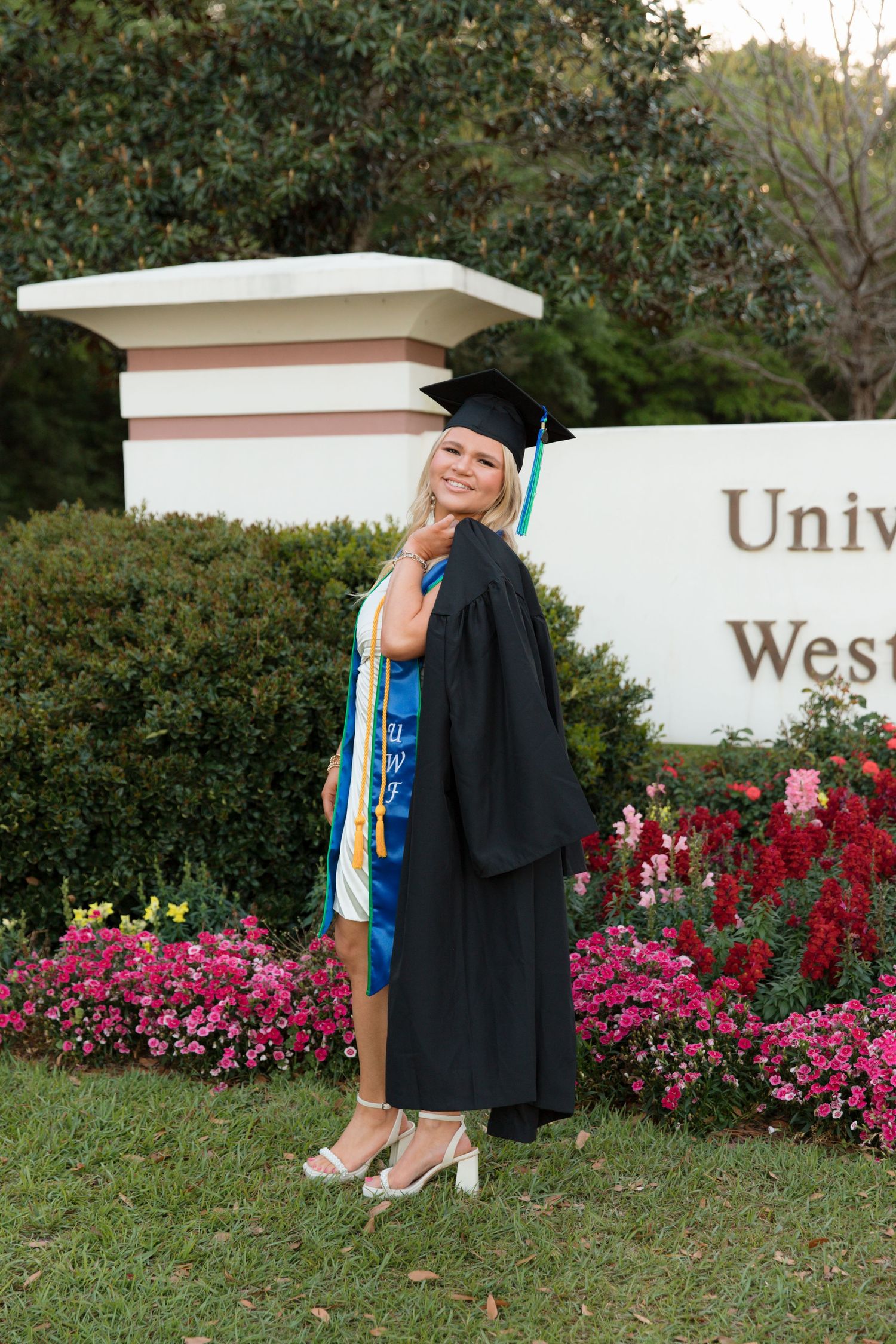 Graduate posing by university sign with blooming pink flowers in foreground.