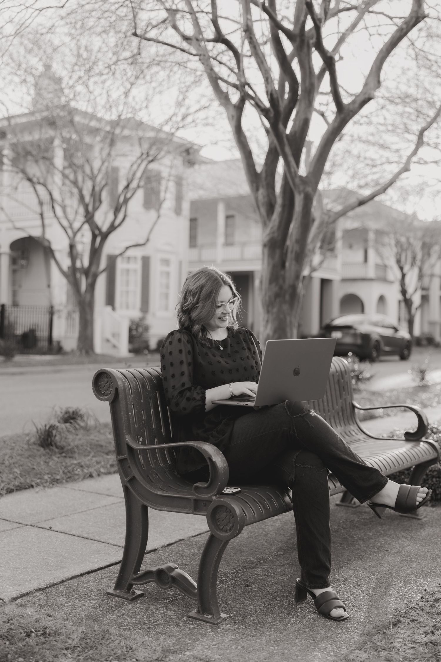 Woman smiling while using laptop on outdoor bench, black and white photo with trees and houses in background.