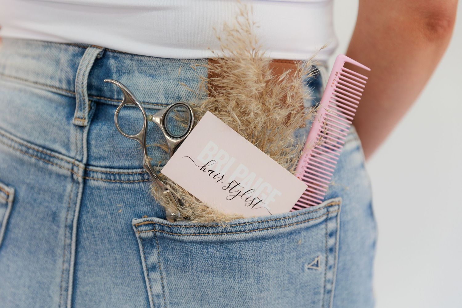 Close-up of styling tools and business card in denim pocket with pampas grass decoration.