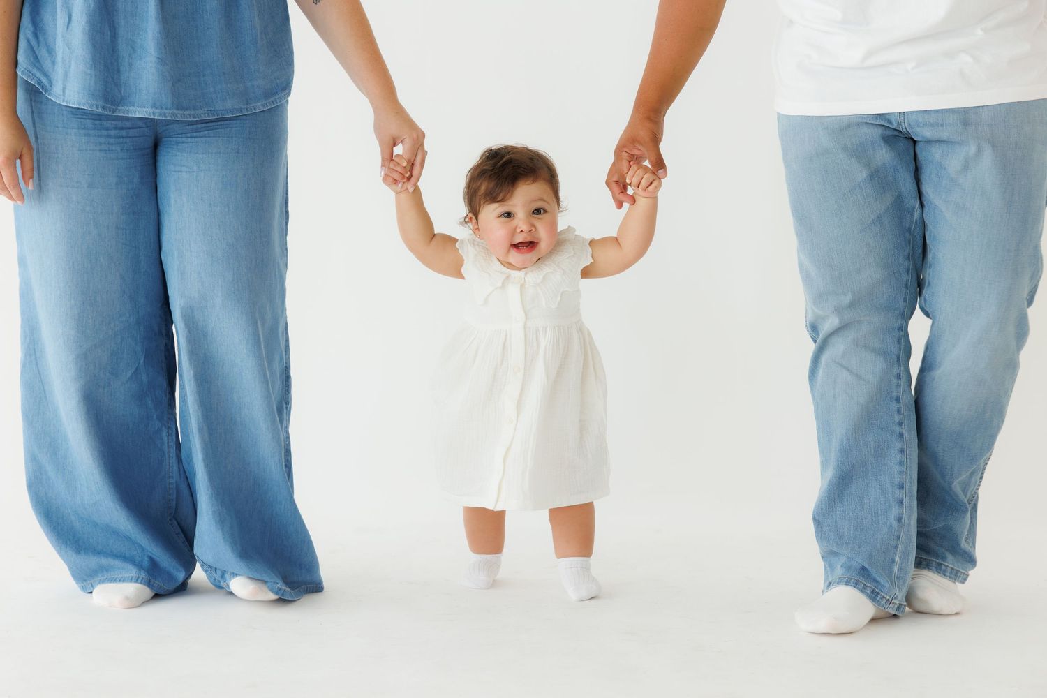 Baby girl in white dress learning to walk while holding hands with two adults in blue jeans on white background.