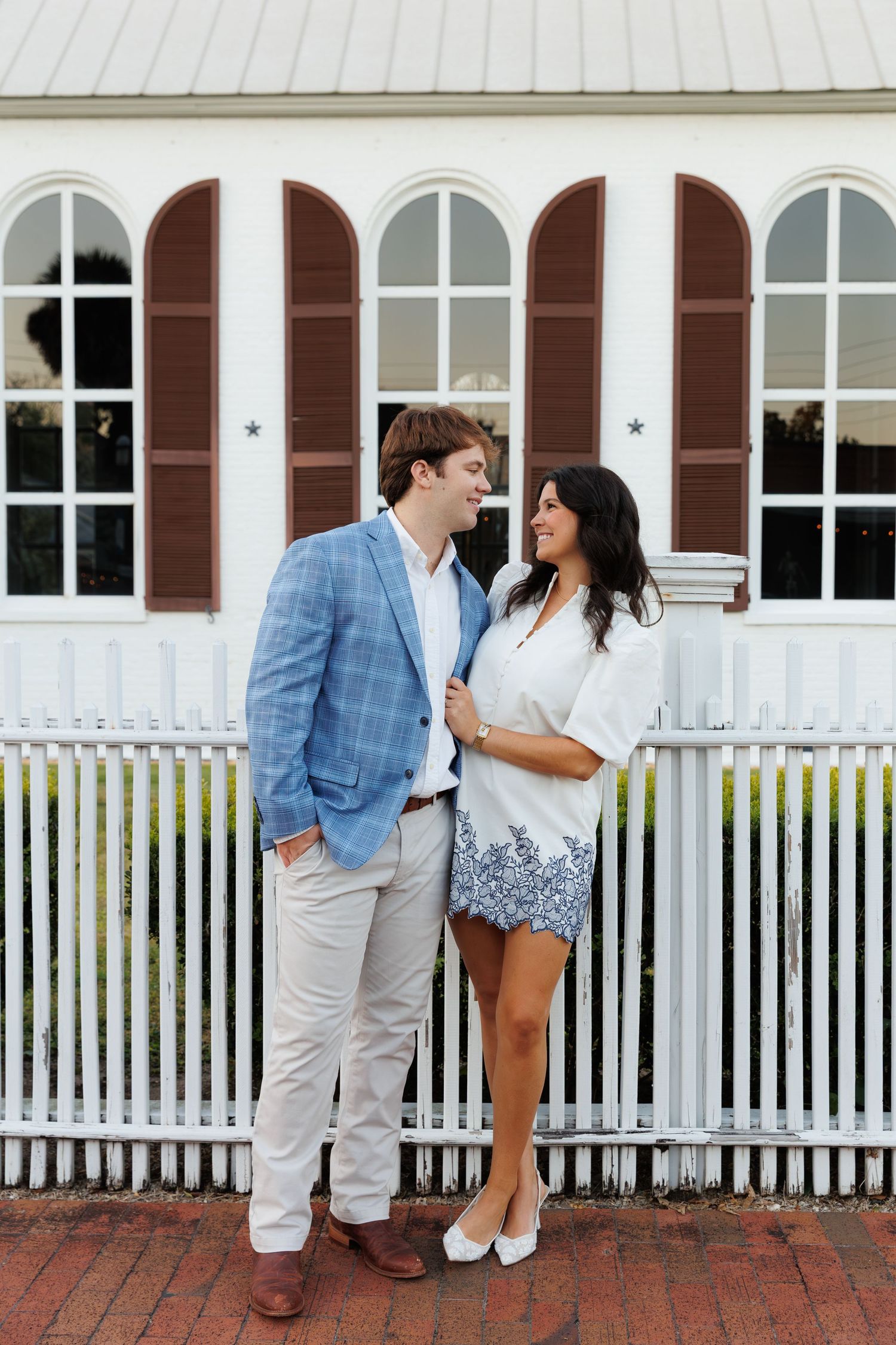 Couple smiling at each other in front of white picket fence and building with arched brown shutters.