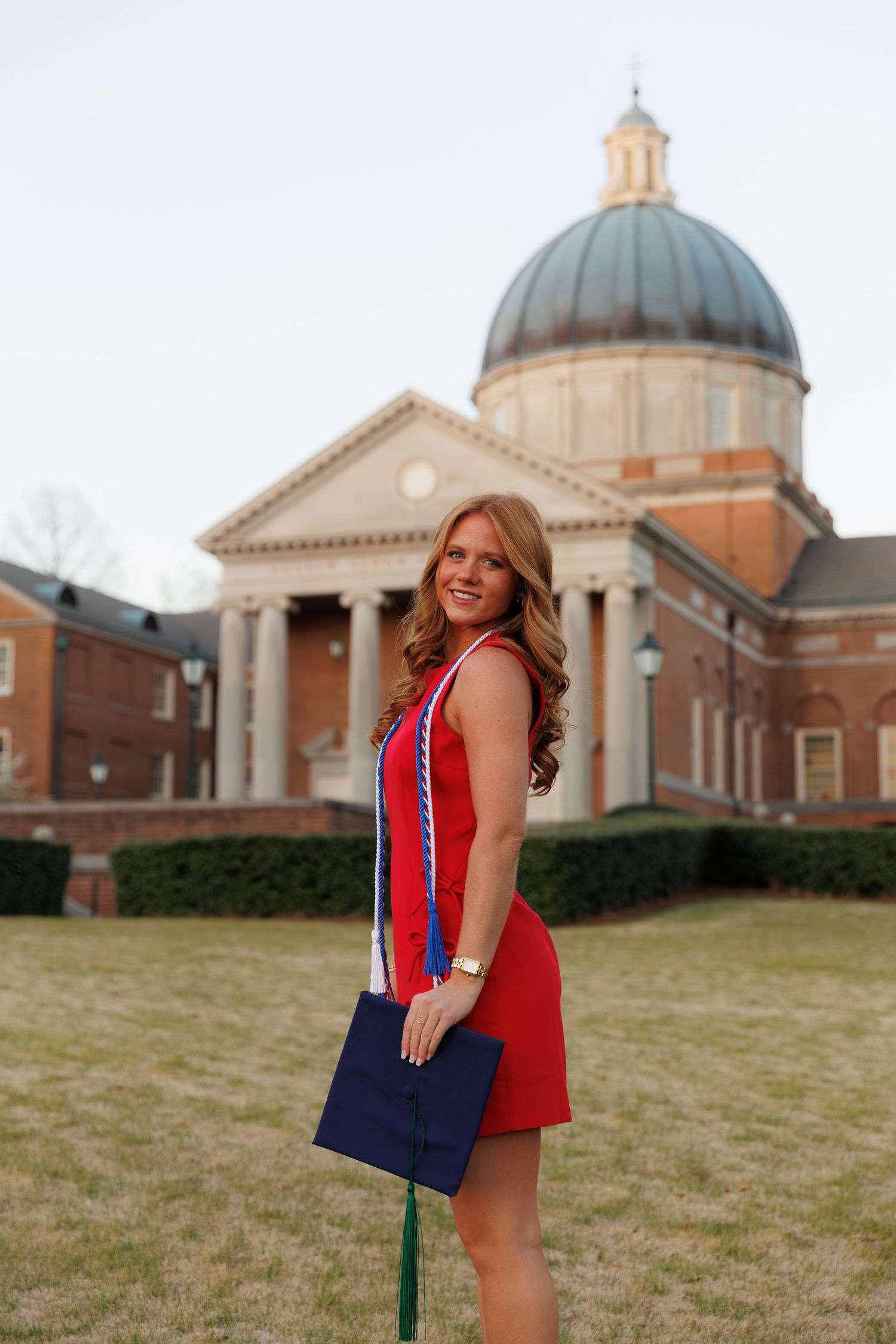 Proud graduate in red dress with honor cords stands before domed university building.