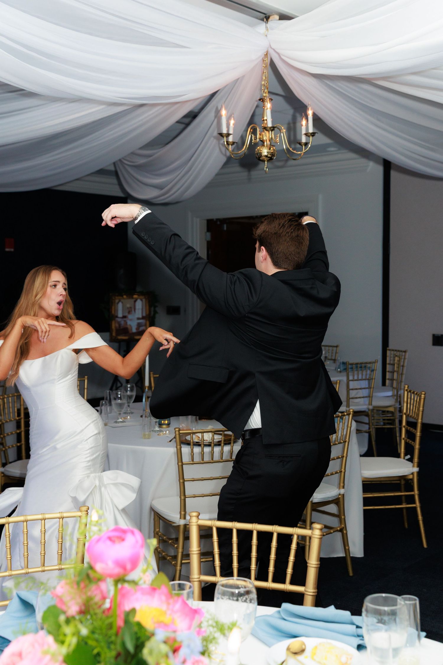 Dancing couple celebrates at wedding reception under draped white fabric ceiling with elegant chandelier and gold chairs.
