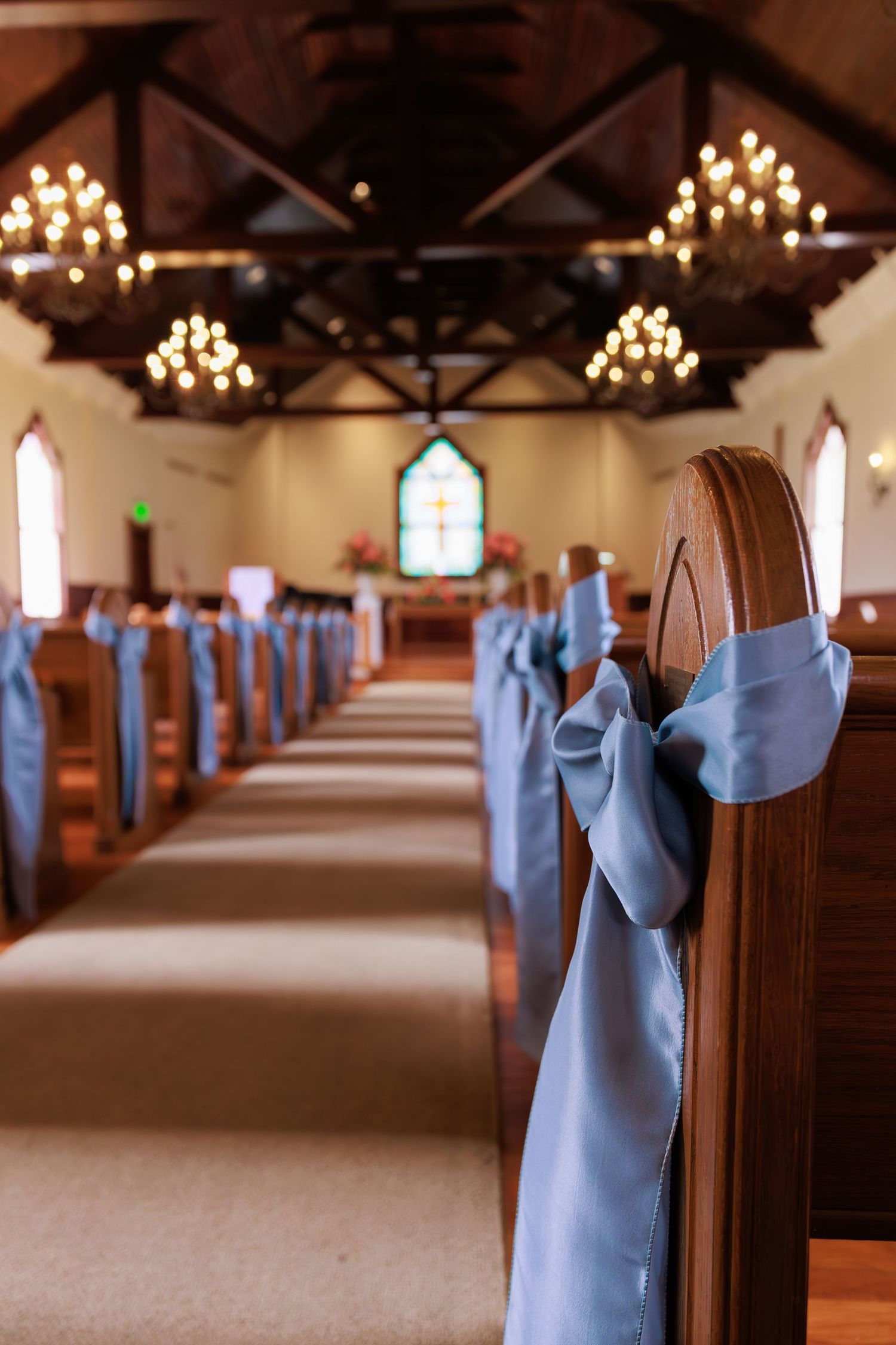 Church aisle decorated with light blue satin bows on wooden pews under wooden beams and chandeliers.