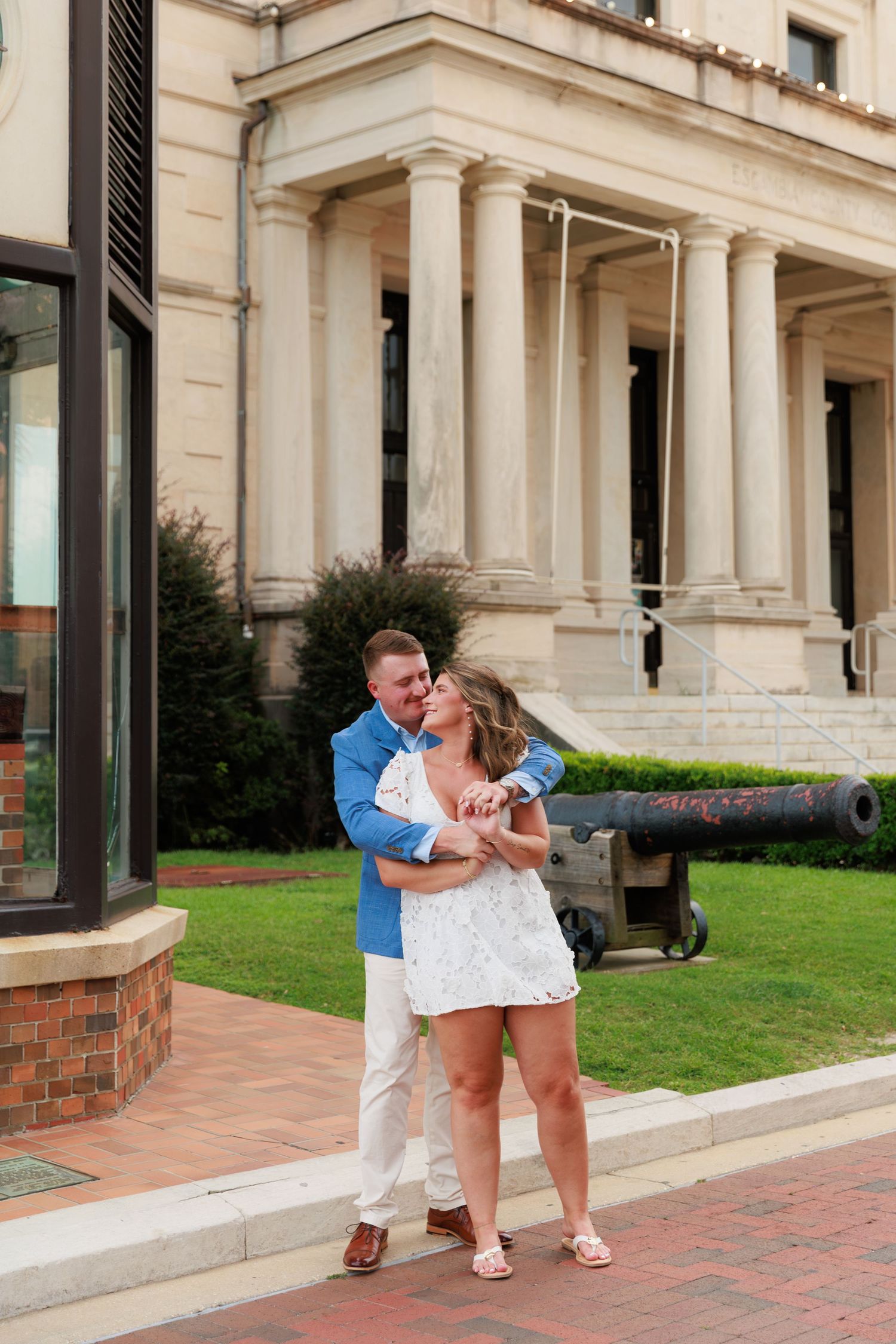 Couple embracing and dancing in front of a grand neoclassical building with columns and a cannon on a brick walkway.