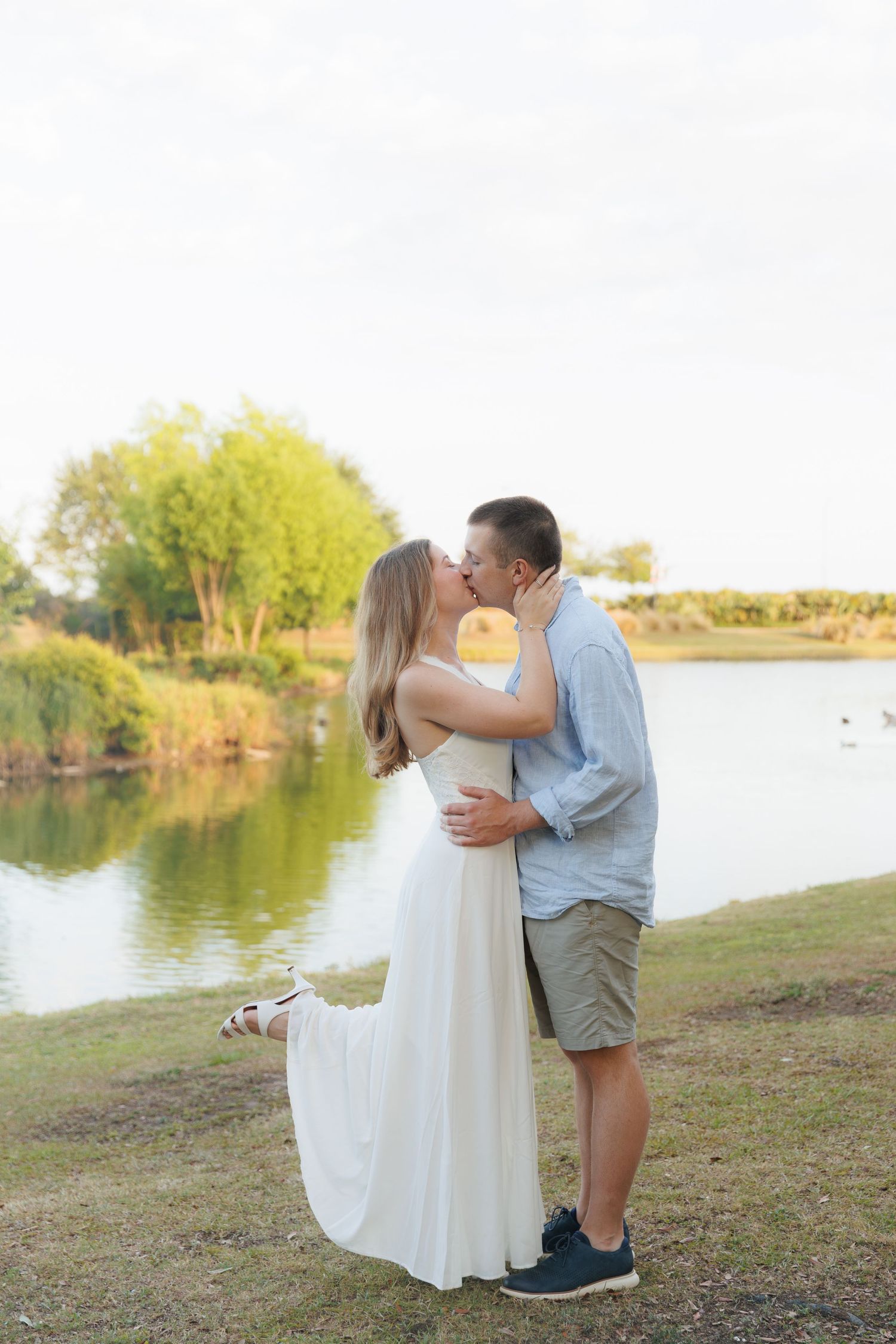 Couple sharing a romantic kiss by a peaceful lake at golden hour, woman in flowing white dress.