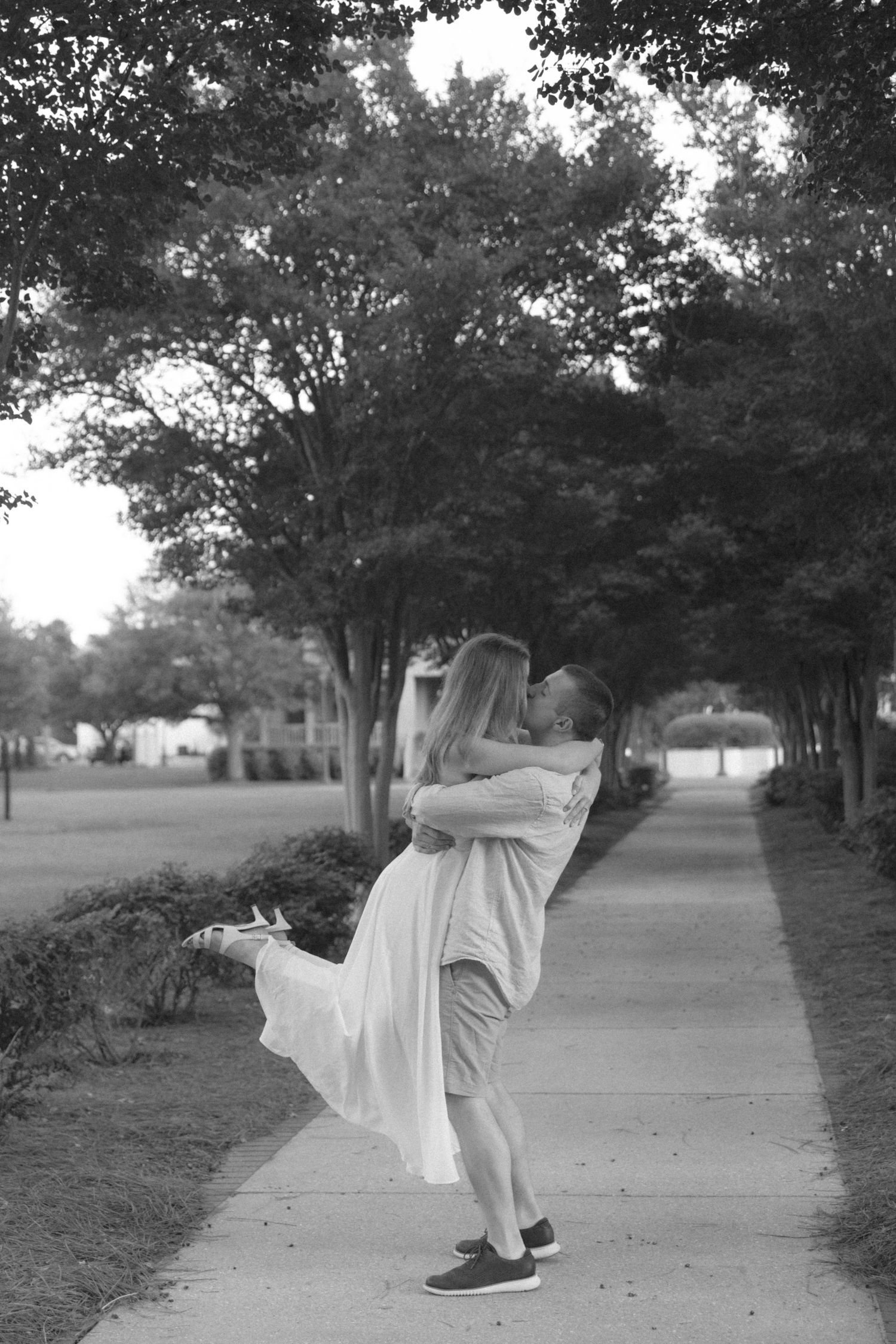 Black and white photo of a man lifting and embracing a woman in a white dress on a tree-lined path.