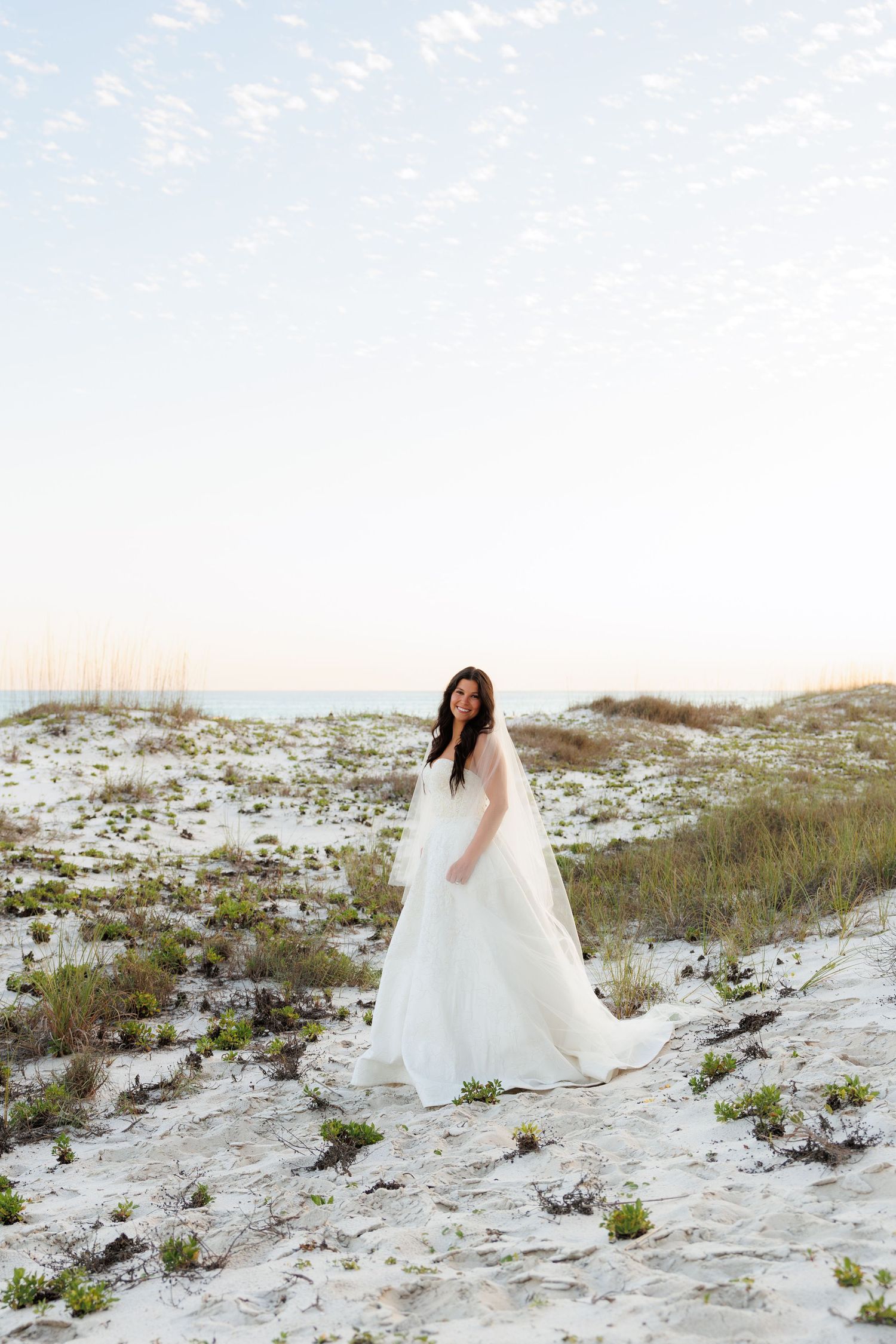 Bride in white wedding gown and cathedral veil stands on sandy beach dunes at sunset.