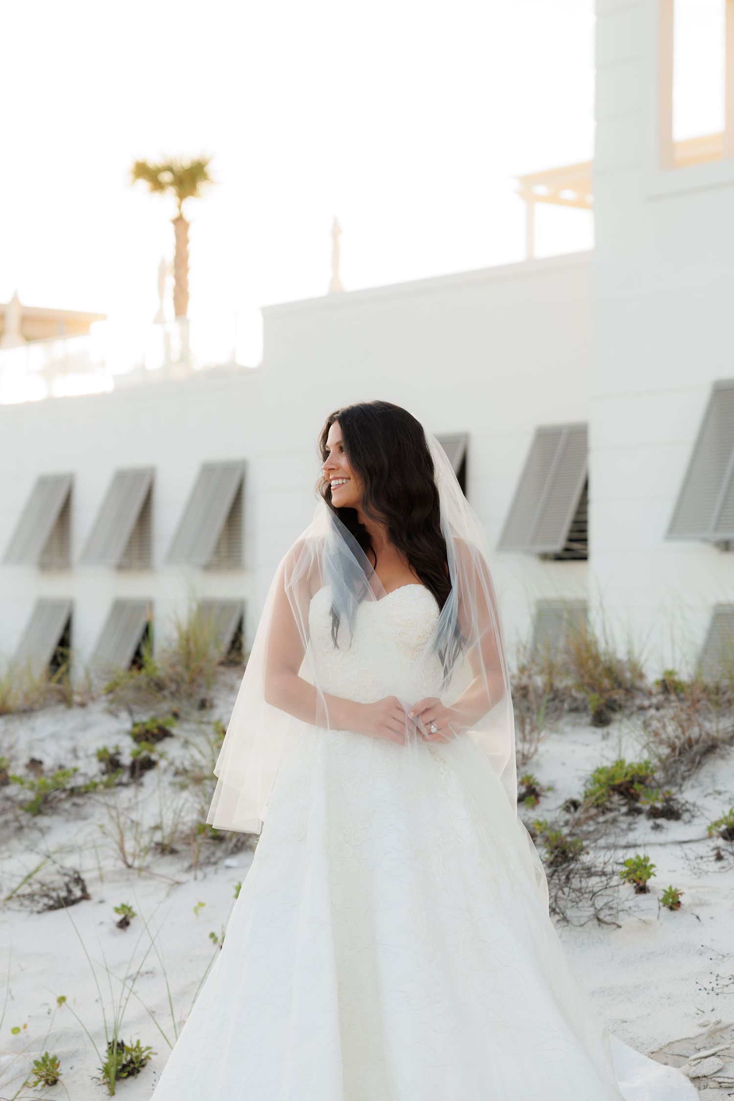 Bride in white strapless gown and veil smiles on sandy beach near white building with palm tree backdrop.