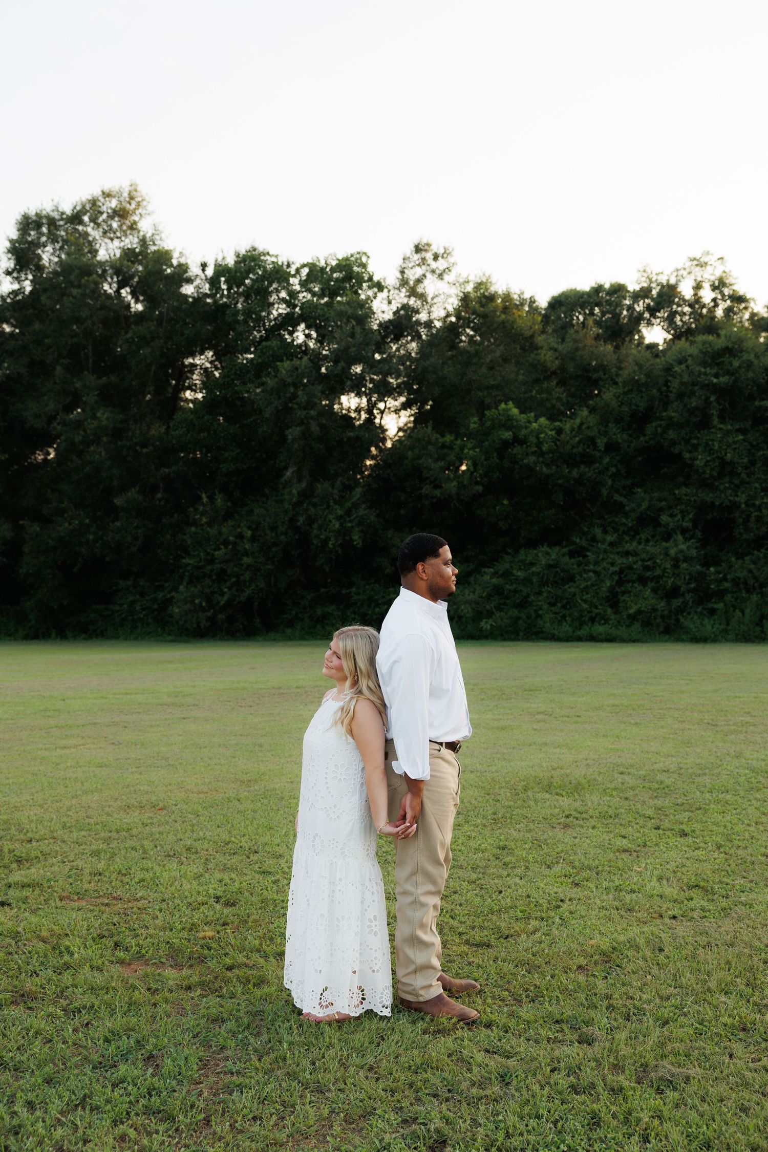 Couple standing back-to-back in open green field at dusk, both wearing white and khaki outfits.