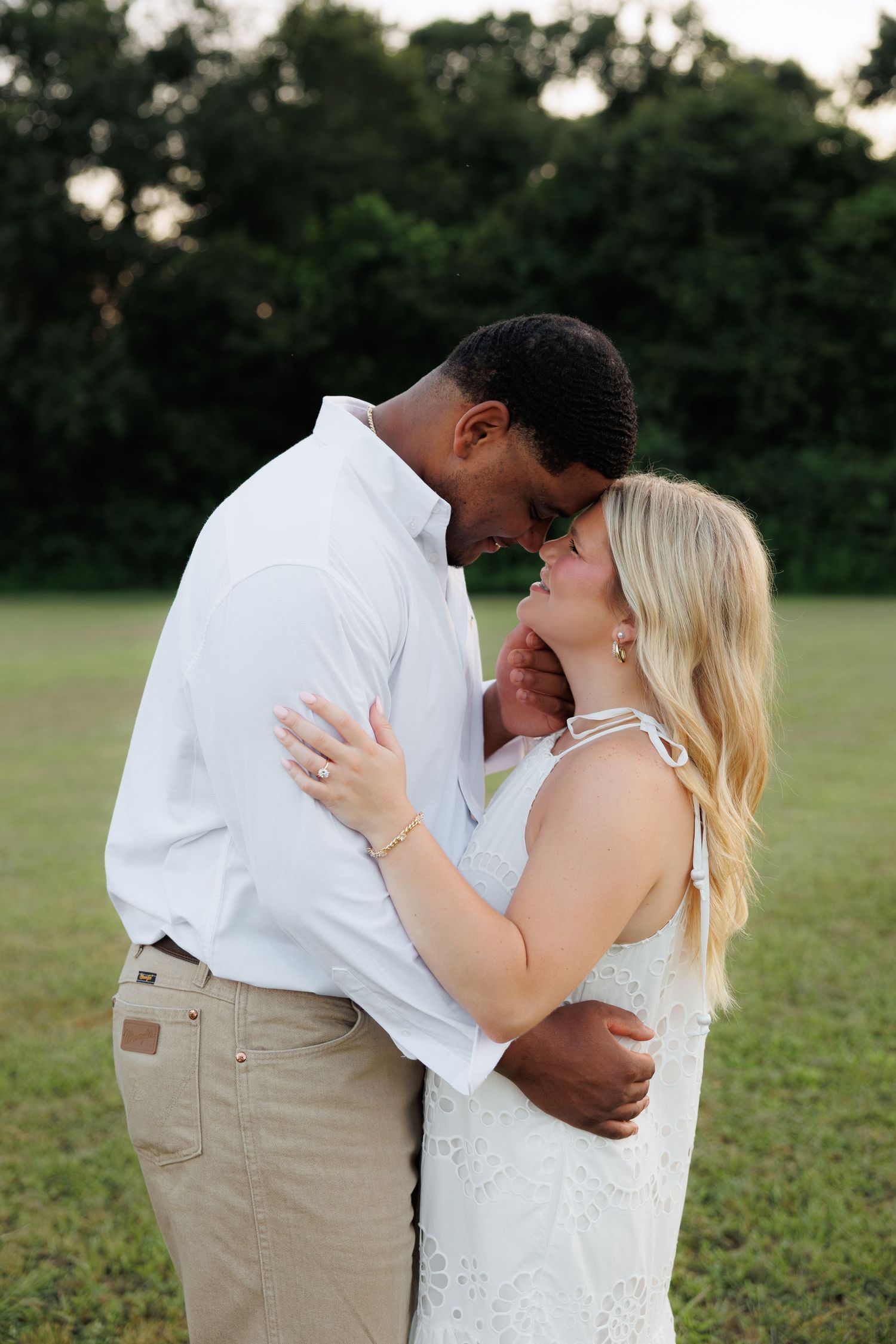 Interracial couple in white outfits sharing a romantic forehead touch in a lush green field at golden hour.