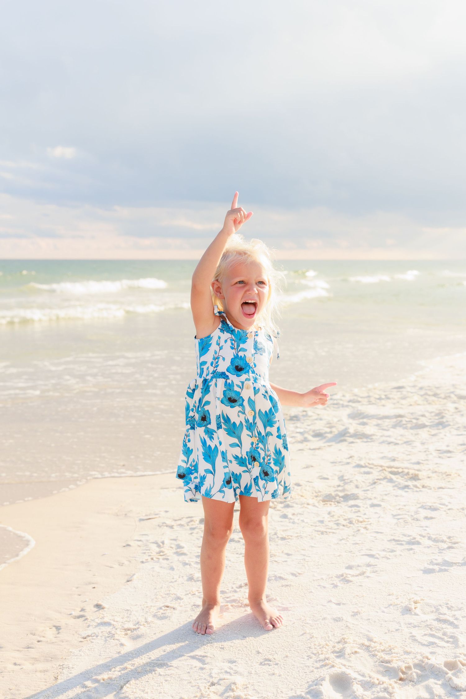 Excited blonde toddler girl in blue floral dress pointing up at the sky on a sandy beach.