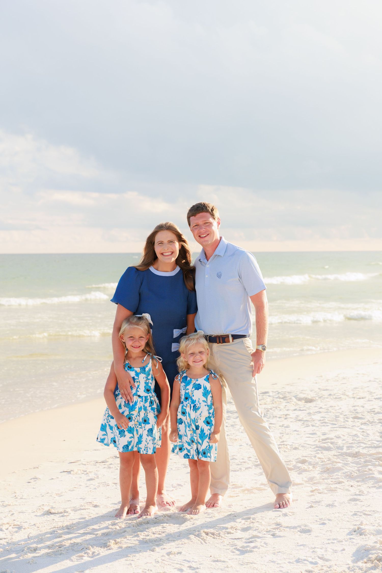 Family of four posing on a white sand beach, parents and two young girls in matching blue floral dresses.
