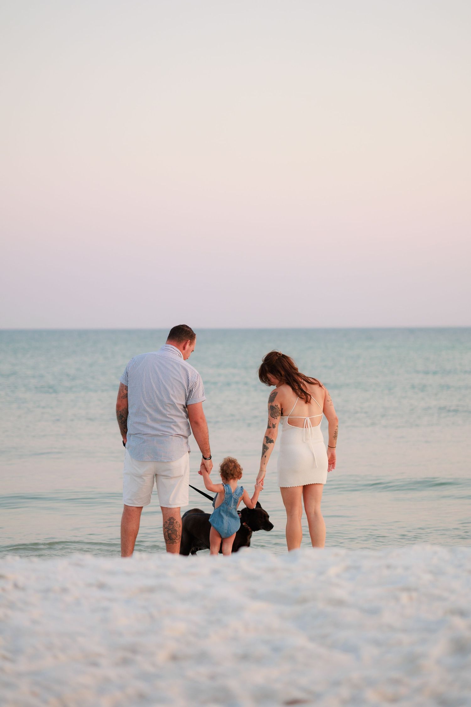 Family with toddler and black dog standing at ocean's edge on a sandy beach at sunset.