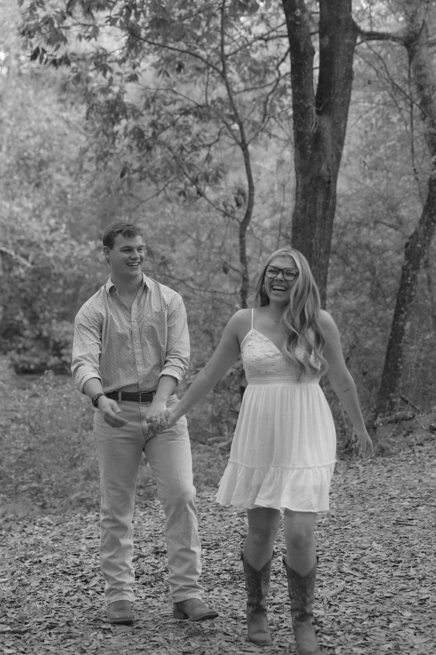 Black and white photo of a smiling couple holding hands in a wooded setting, woman in white dress and boots.