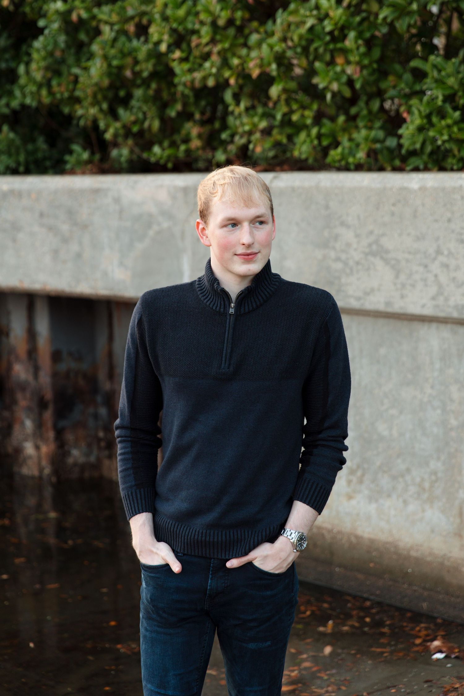 Young man in black zip-neck sweater and jeans standing outdoors near a concrete wall with green shrubs.