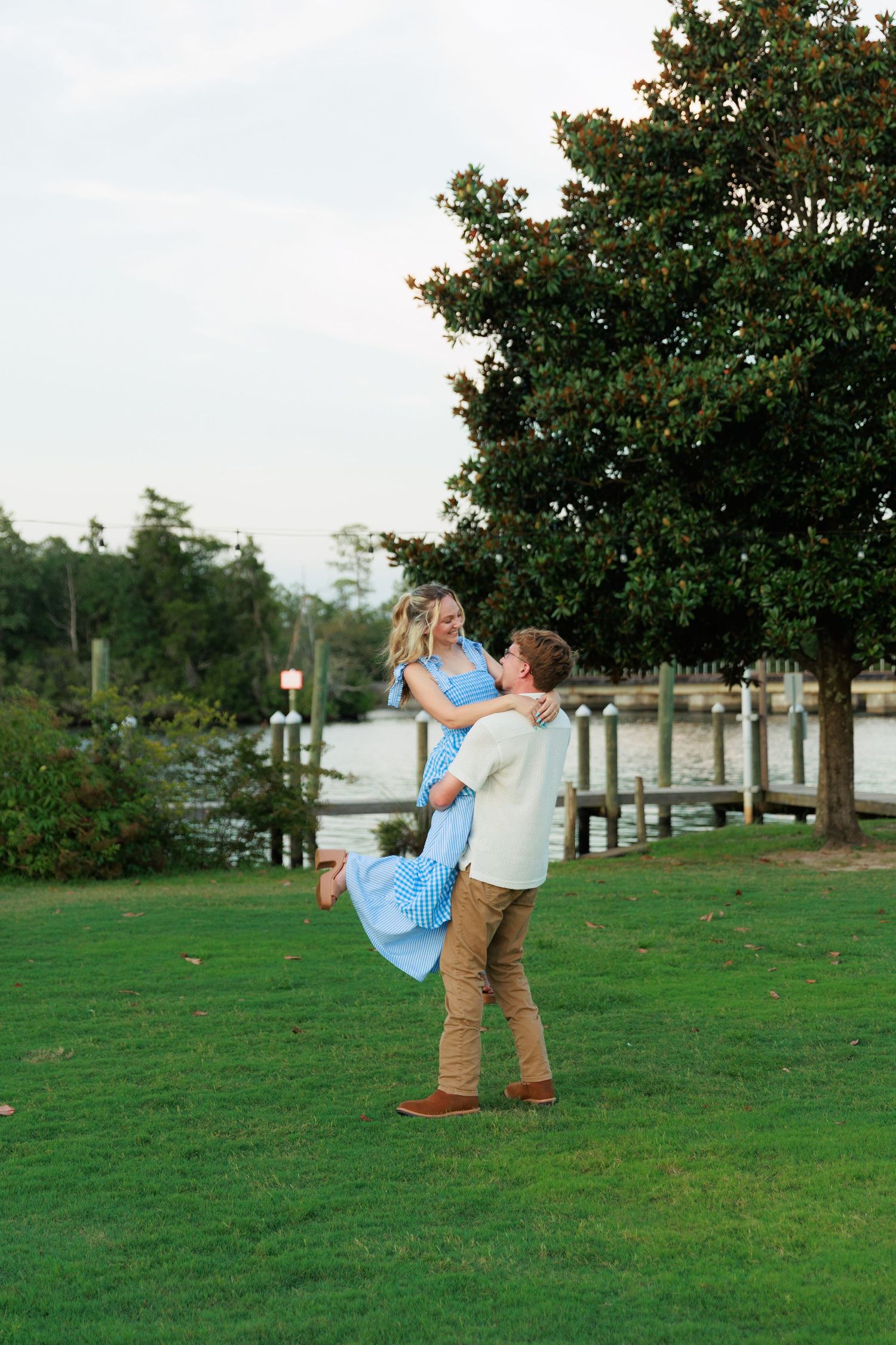 A man joyfully lifts his partner in a blue gingham dress on a green lawn near a scenic waterfront at dusk.
